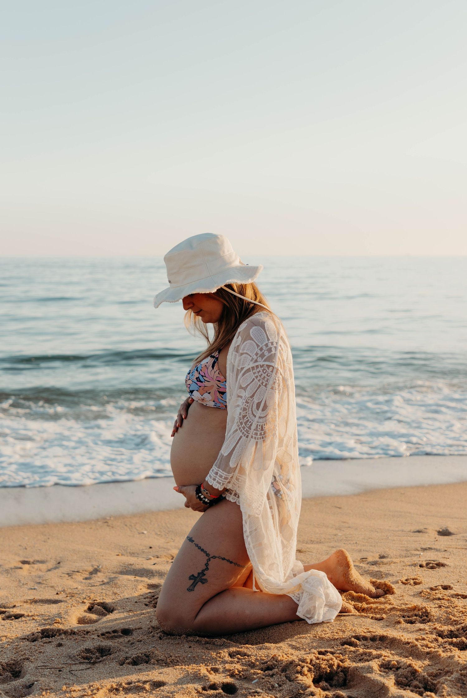 Pregnant woman in white hat and lace kimono kneels on sandy beach at sunset.