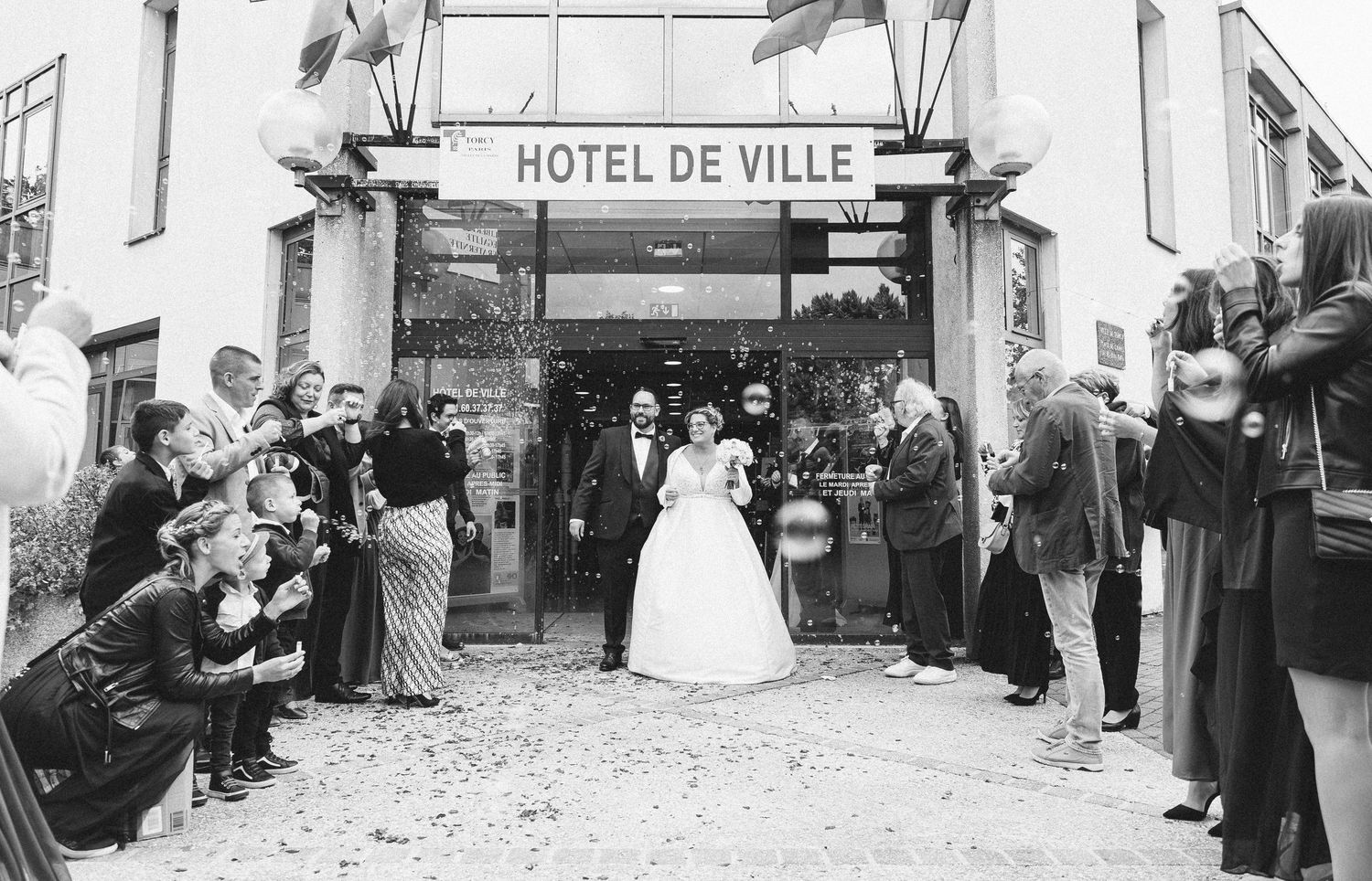A vintage black and white wedding photo outside Hotel de Ville with guests throwing confetti.