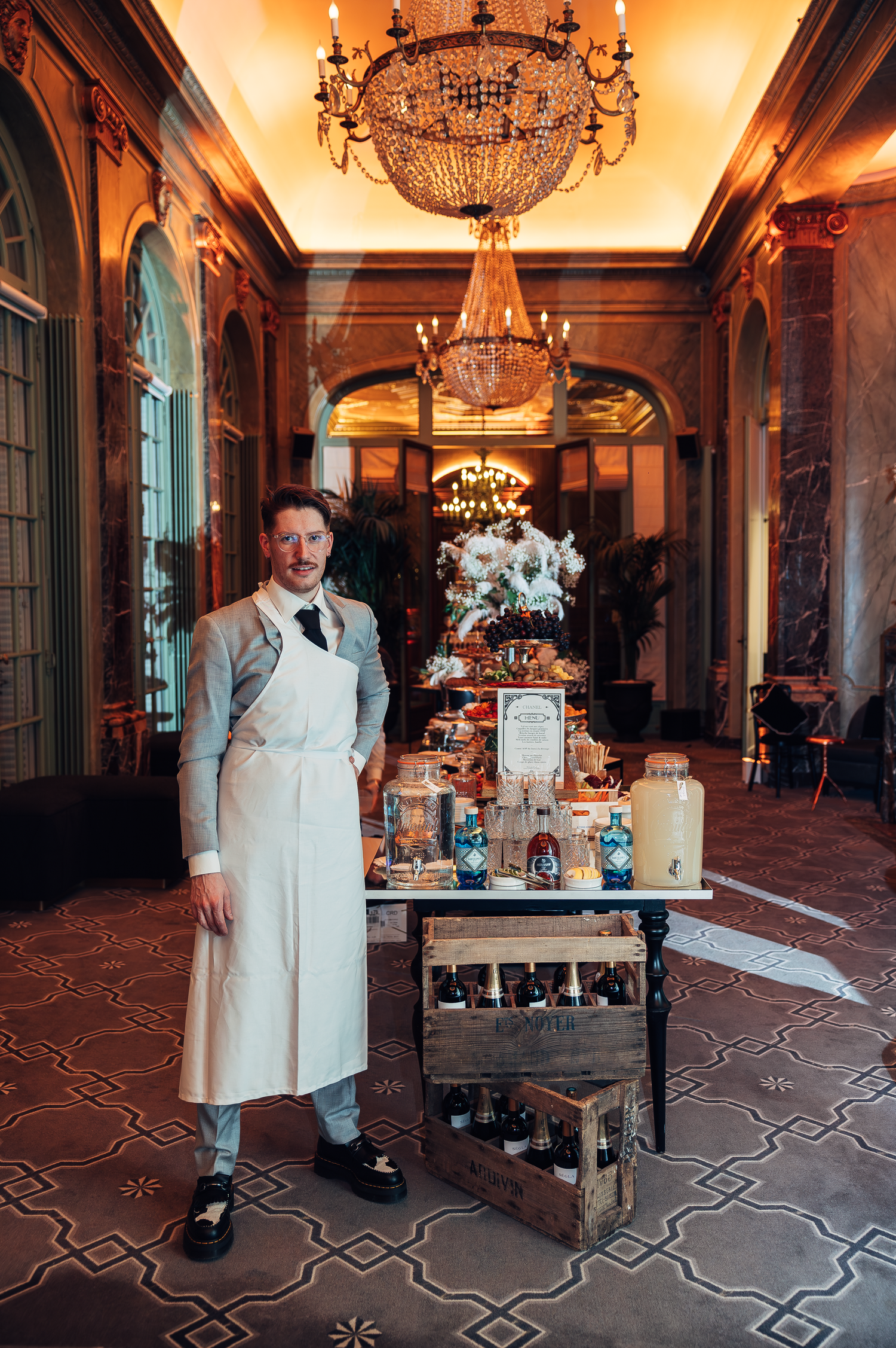 Staff member in vintage uniform stands beside antique beverage cart under crystal chandeliers.