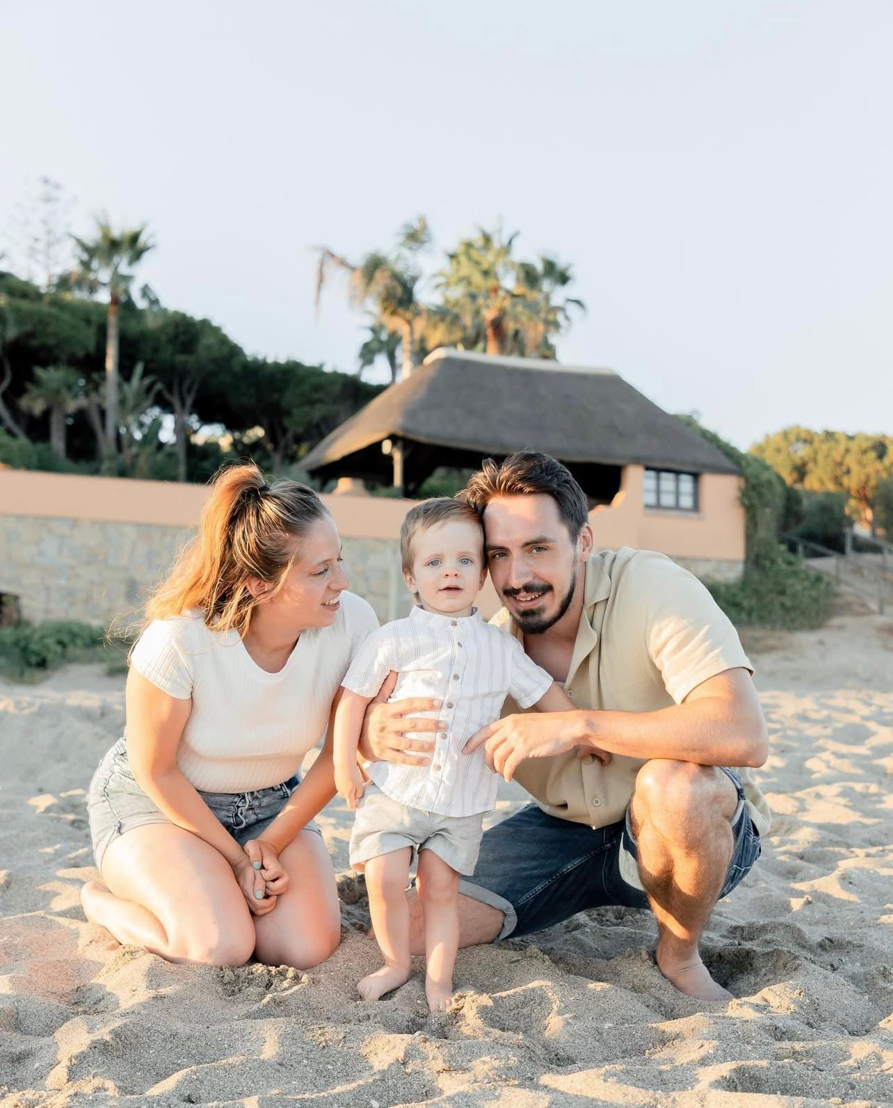 Family enjoying a sunset moment together on a sandy beach with palm trees in the background.