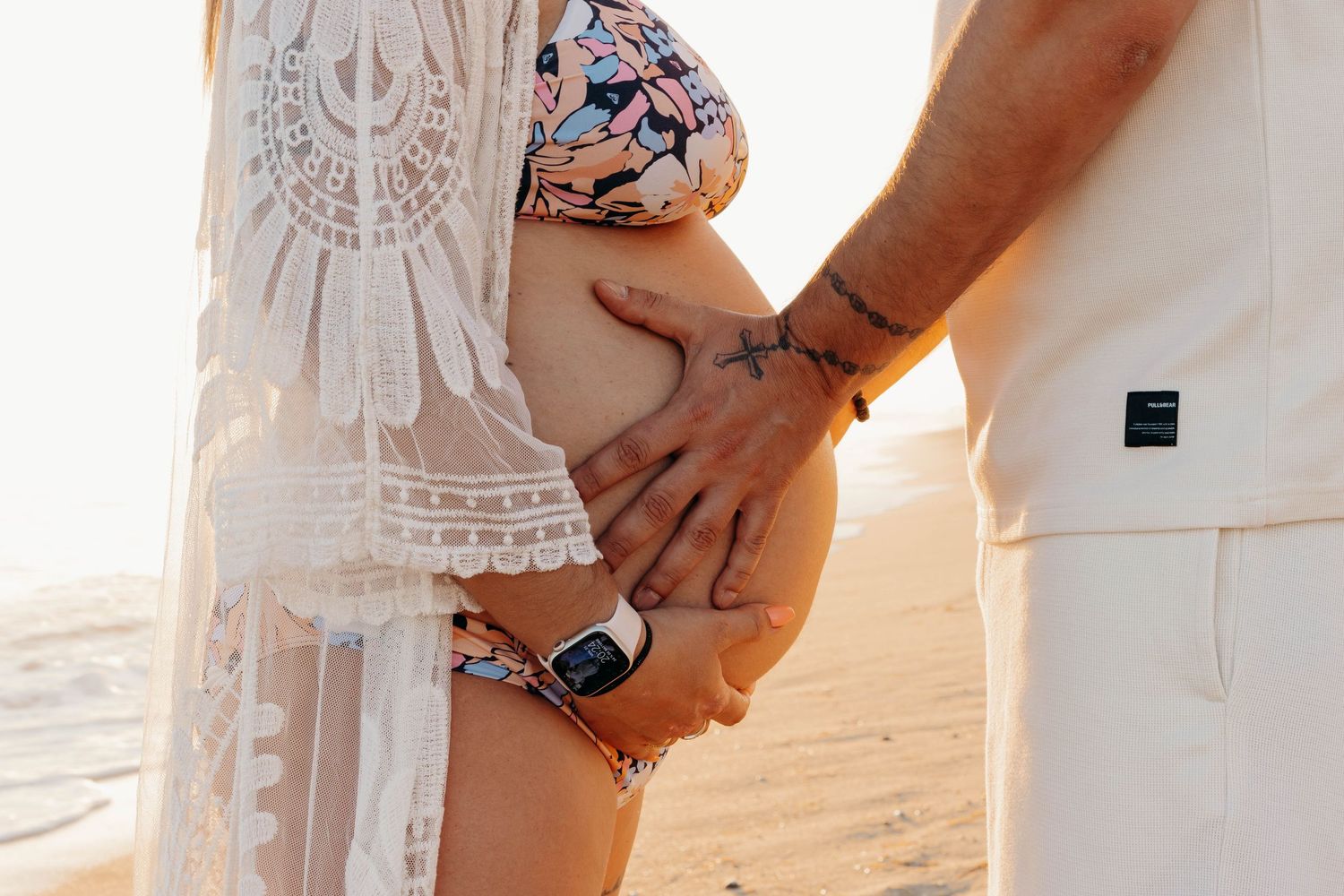 Close-up of hands gently cradling a pregnant belly with lace dress detail at sunset.