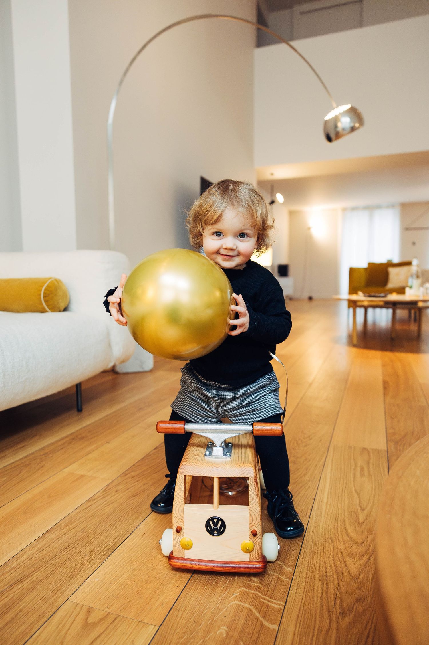 Small child plays with a golden balloon while sitting on a toy VW bus on hardwood floors in a modern living room.