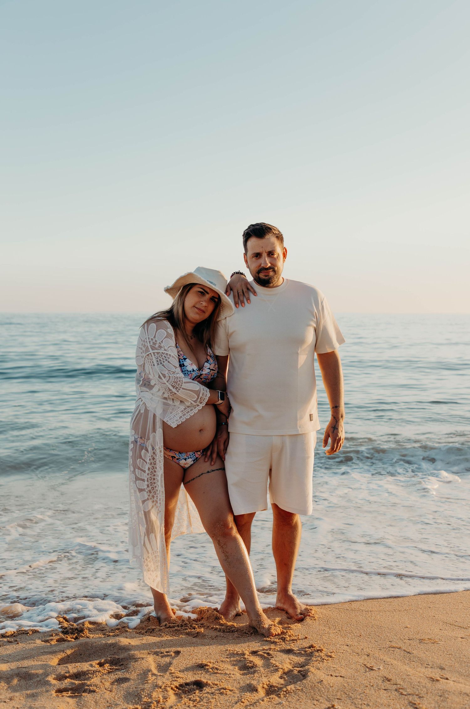 A couple stands together on a beach at sunset with ocean waves in the background.