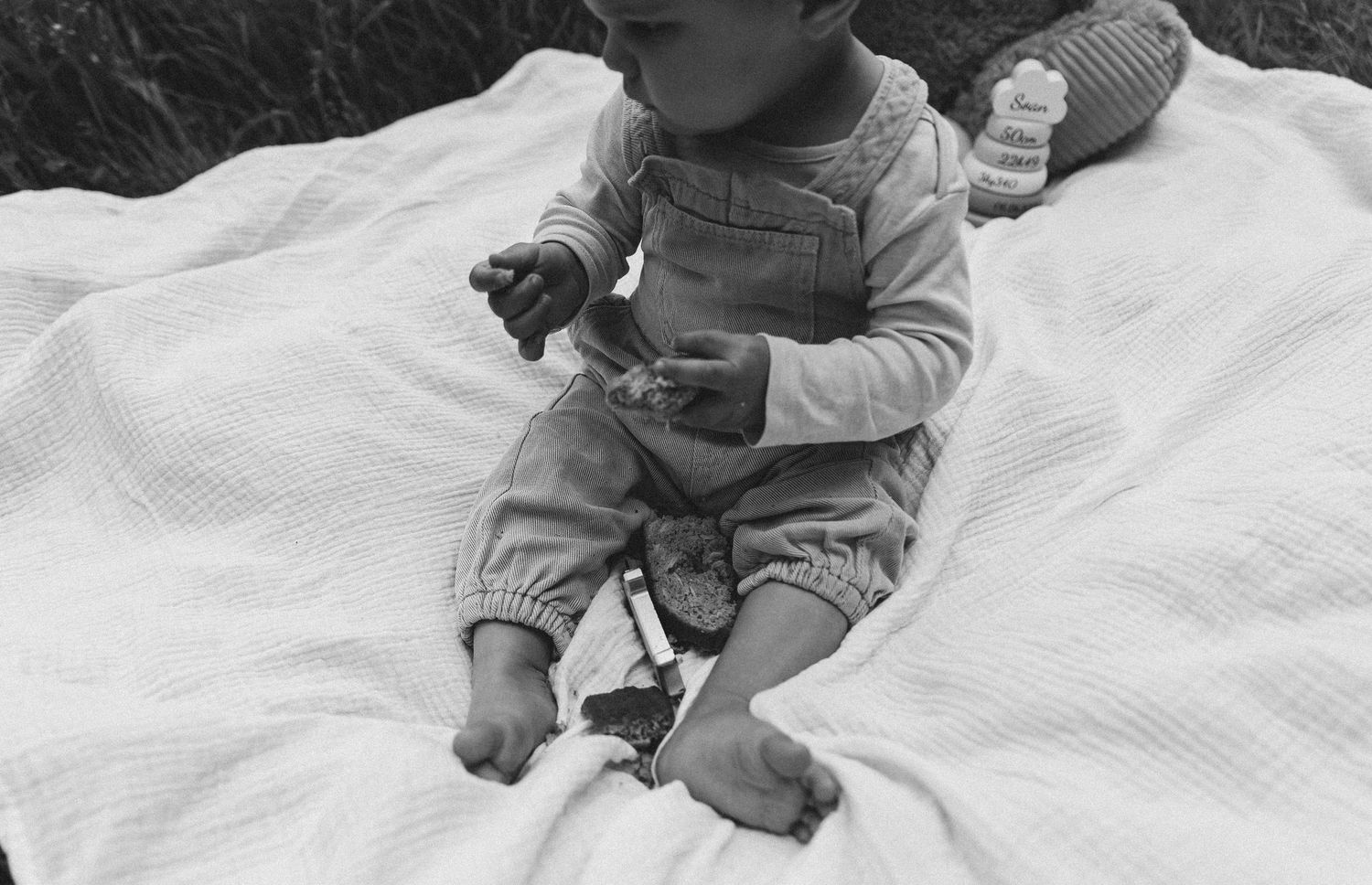 Black and white image of baby in overalls sitting on blanket outdoors.