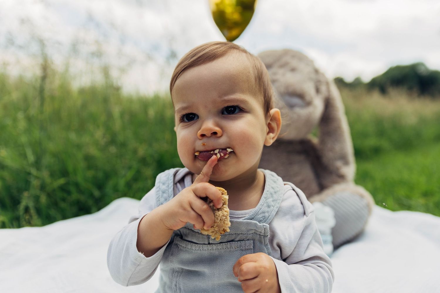 Baby in light blue overalls smiles while sitting on white blanket with stuffed bunny.