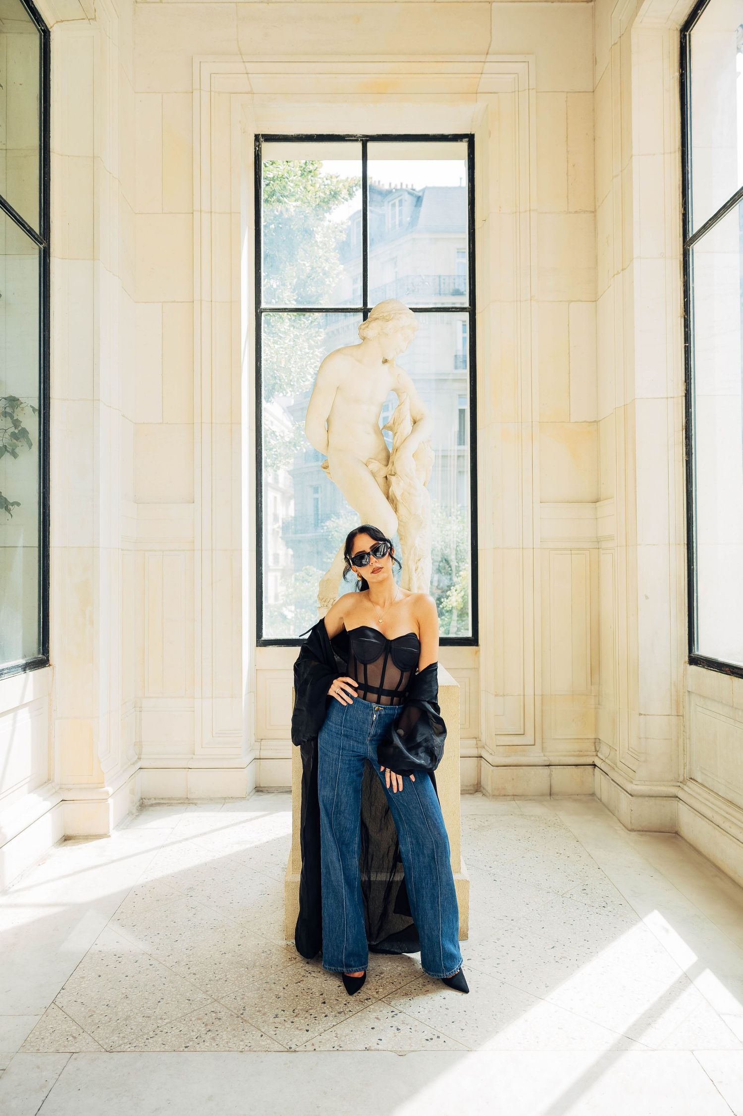Dramatic fashion pose in a sunlit room with black corset top and flowing coat.