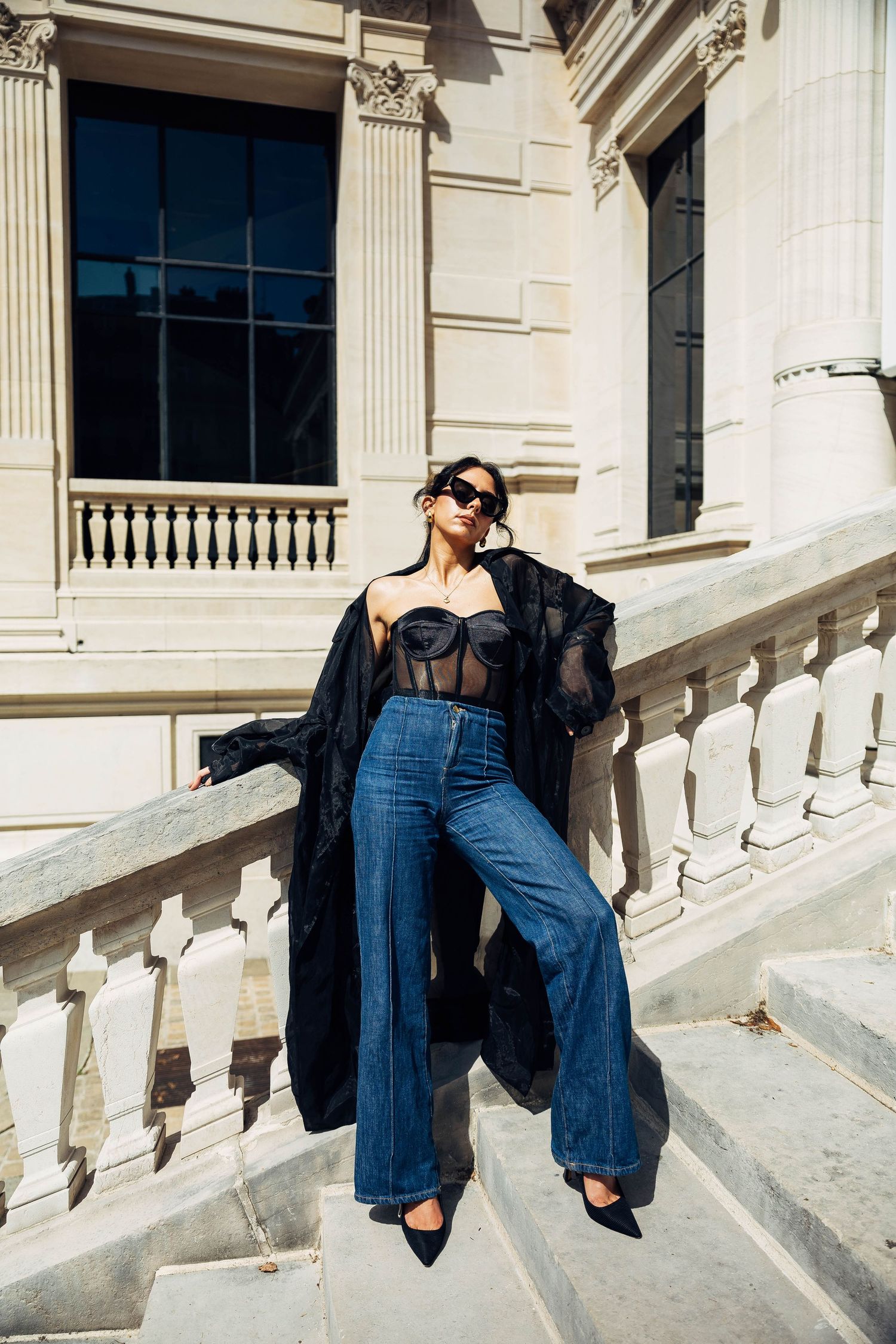 Editorial fashion shot on marble steps with sheer black coat and wide-leg jeans.
