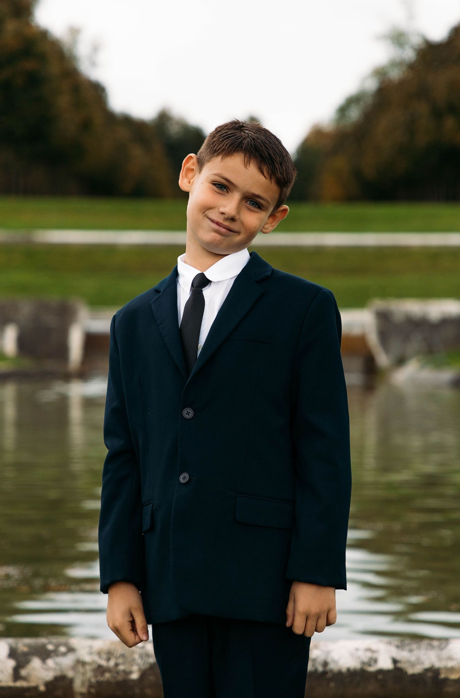 A formal portrait of a young boy in a suit near a lake.