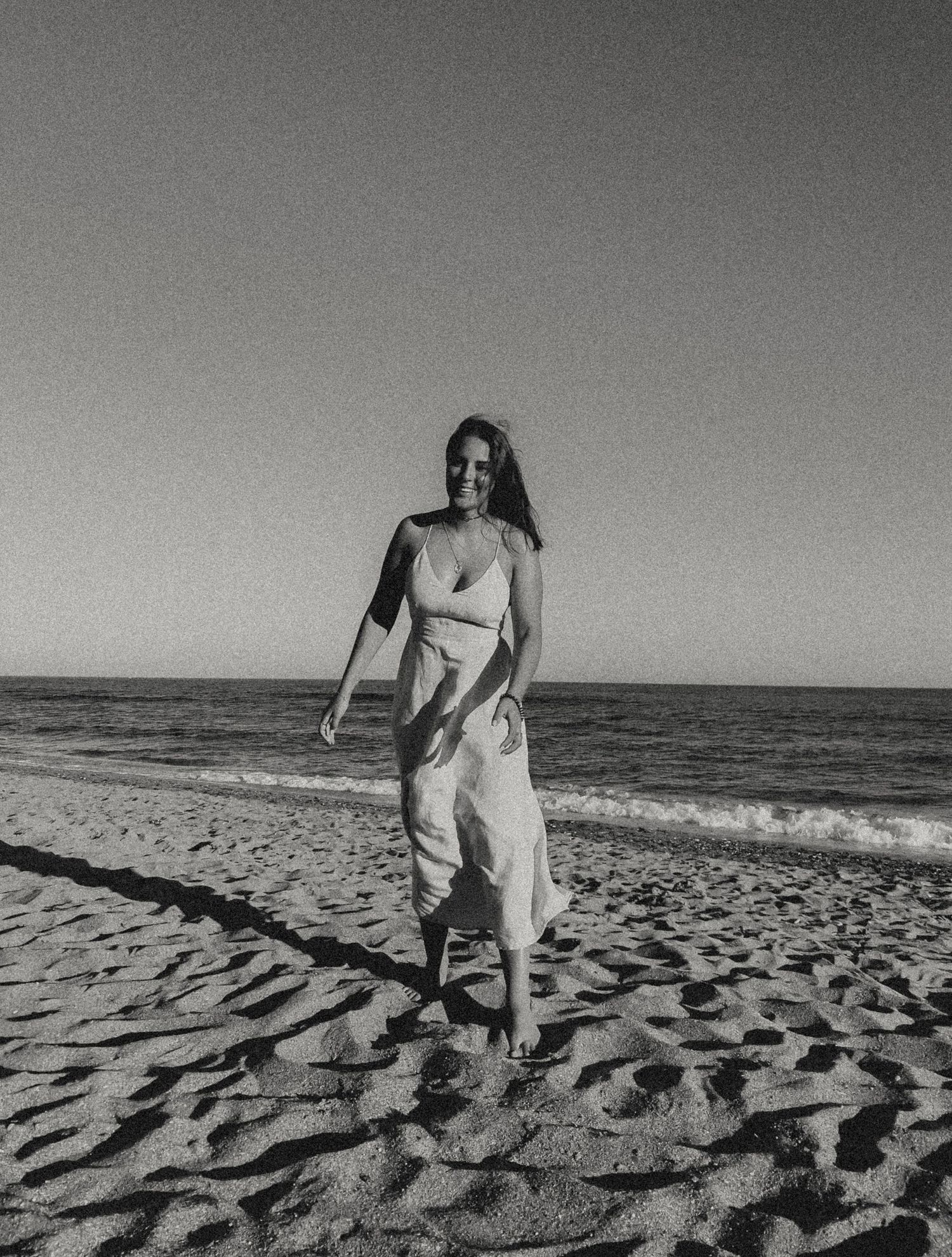A black and white photo of someone walking along a beach in a flowing dress at sunset.