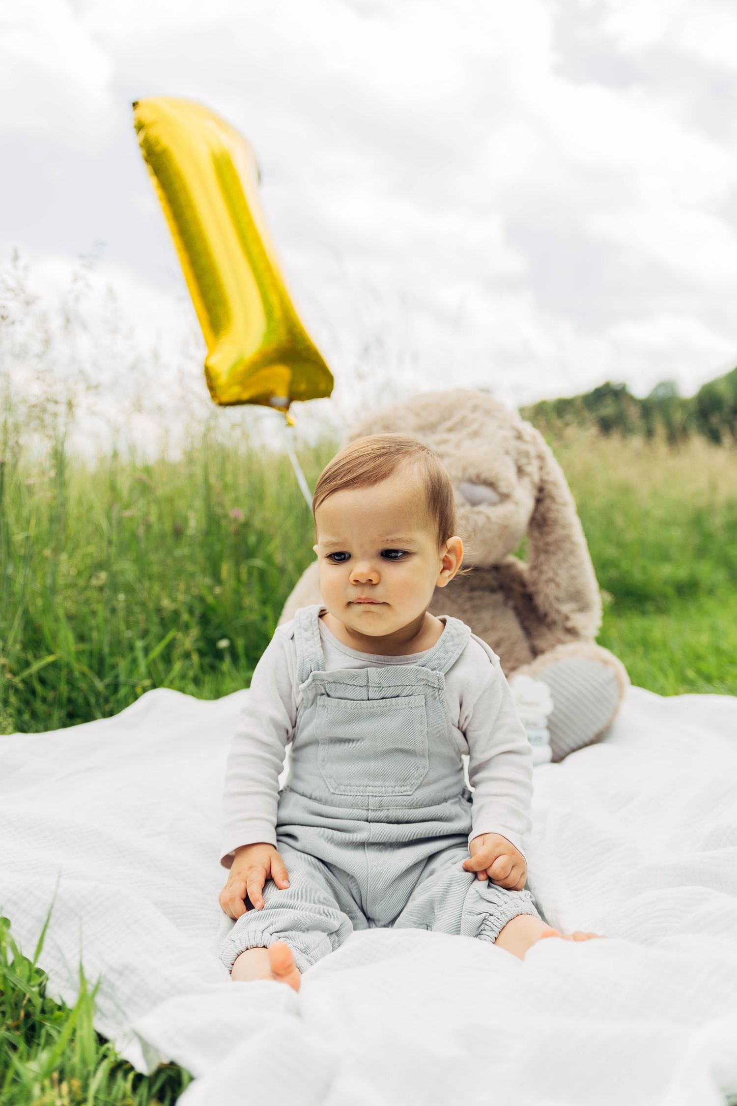 Baby in grey overalls sits on white blanket with yellow balloon and plush bunny.