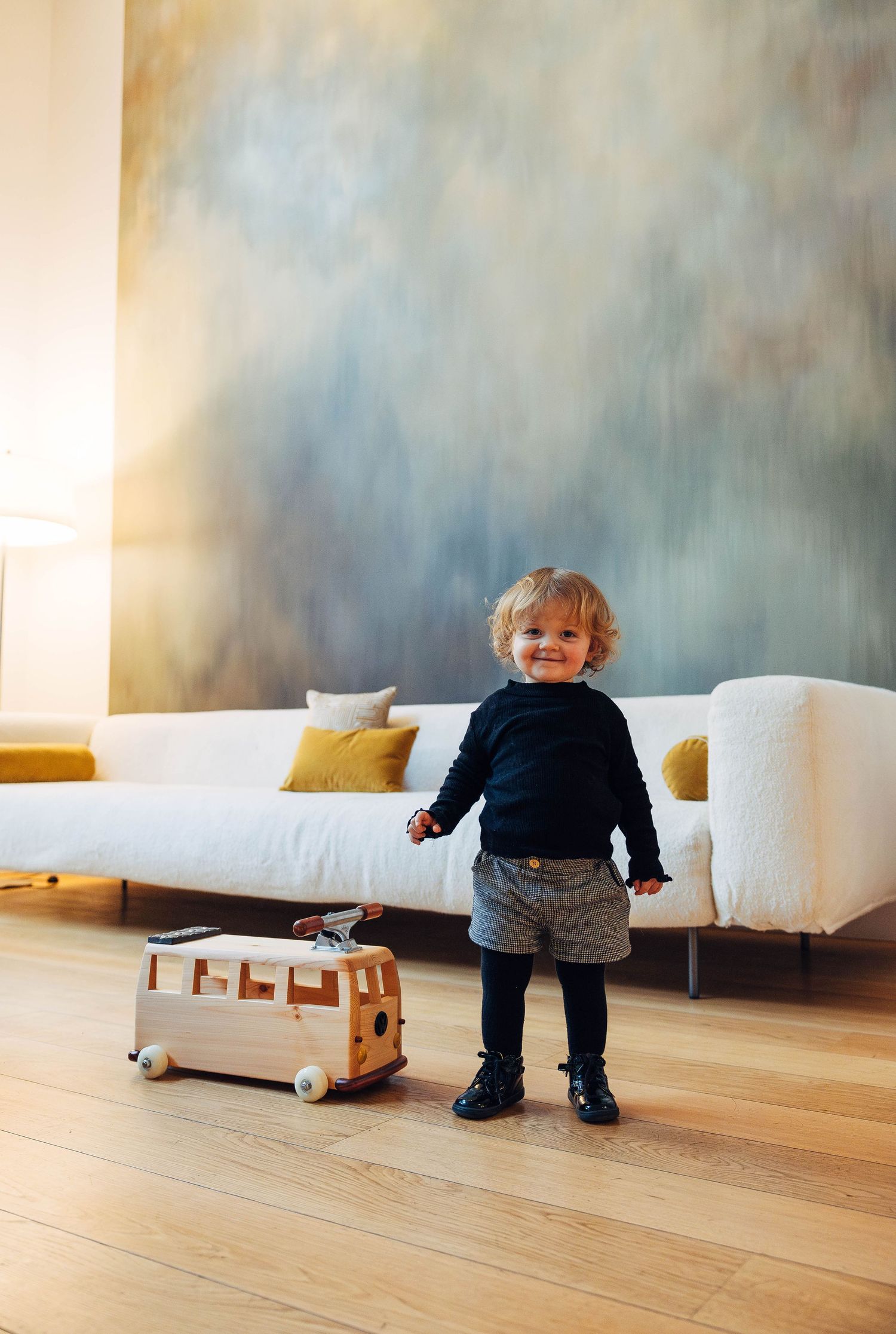A small child wearing dark clothes walks away from a white couch on hardwood flooring in a minimalist room.