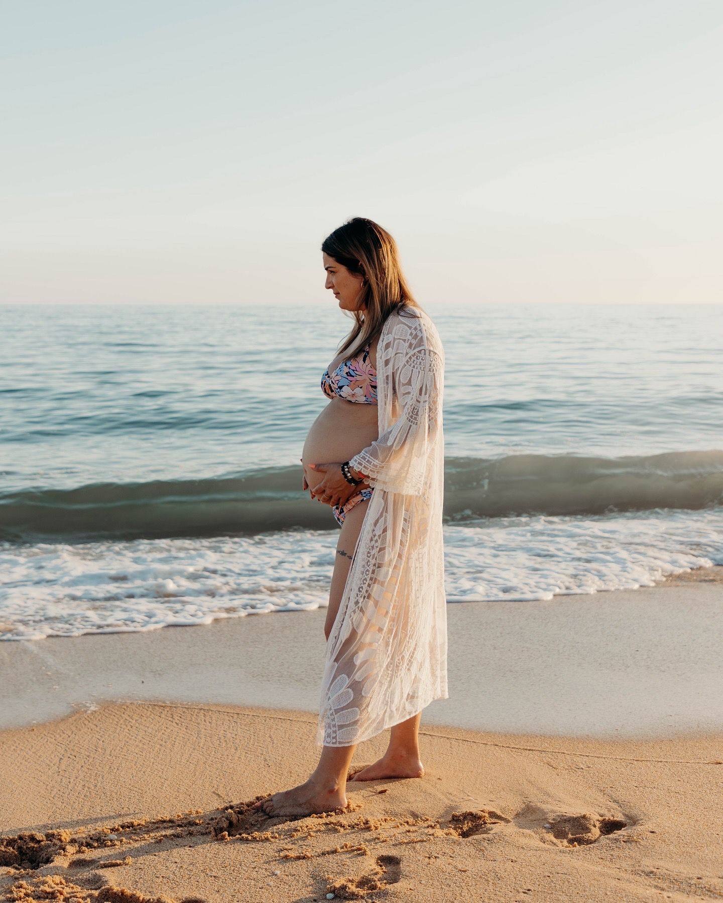 Expectant mother in a lace cover-up poses on beach at sunset.