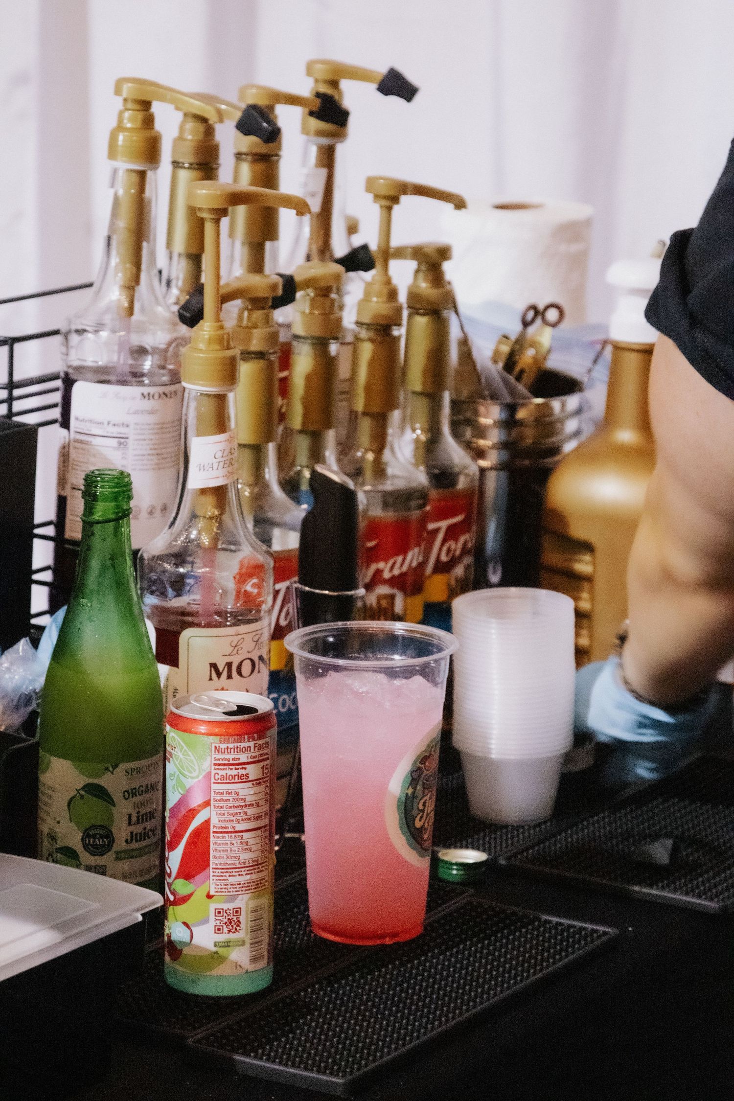 Collection of drink bottles and syrups arranged on black bar mat.