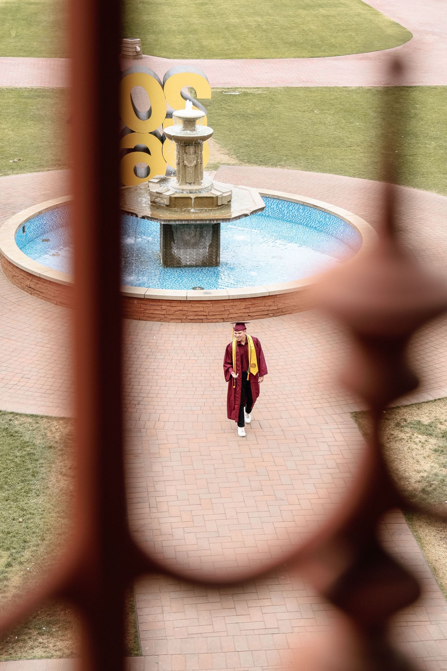 Graduate in maroon cap and gown stands near a decorative fountain on a brick plaza, viewed through ornate iron railing.