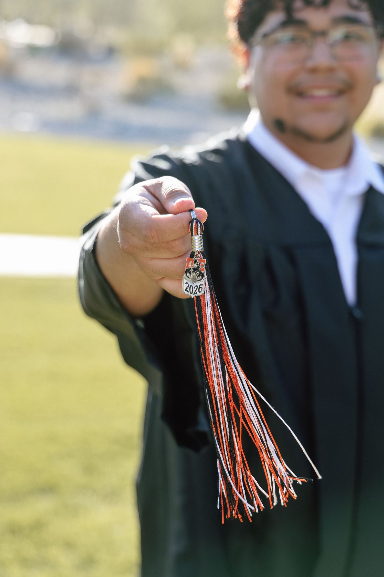 Graduate in black gown holding red and orange tassel with 2020 charm toward camera, smiling outdoors.