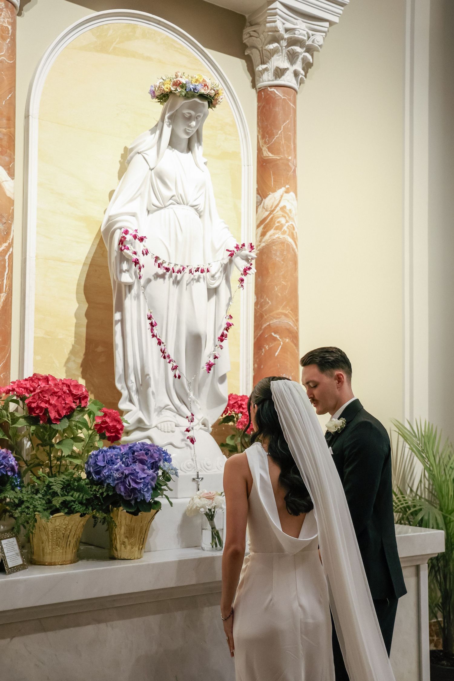 Bride and groom pray before Virgin Mary statue adorned with flowers during Catholic wedding ceremony.