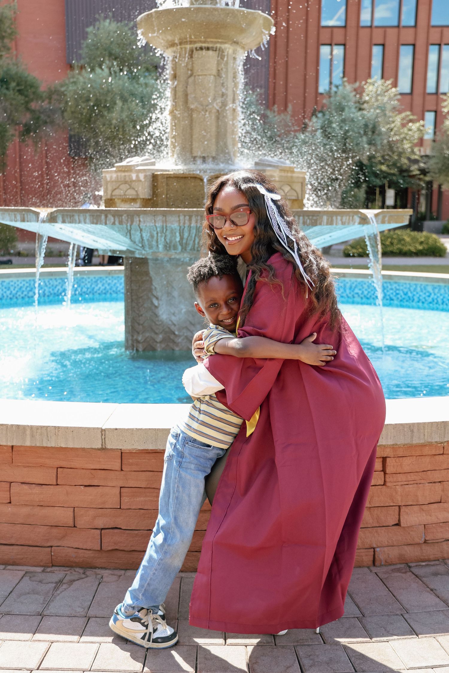 A graduate in maroon gown hugs a young boy by a fountain, celebrating her graduation outdoors.