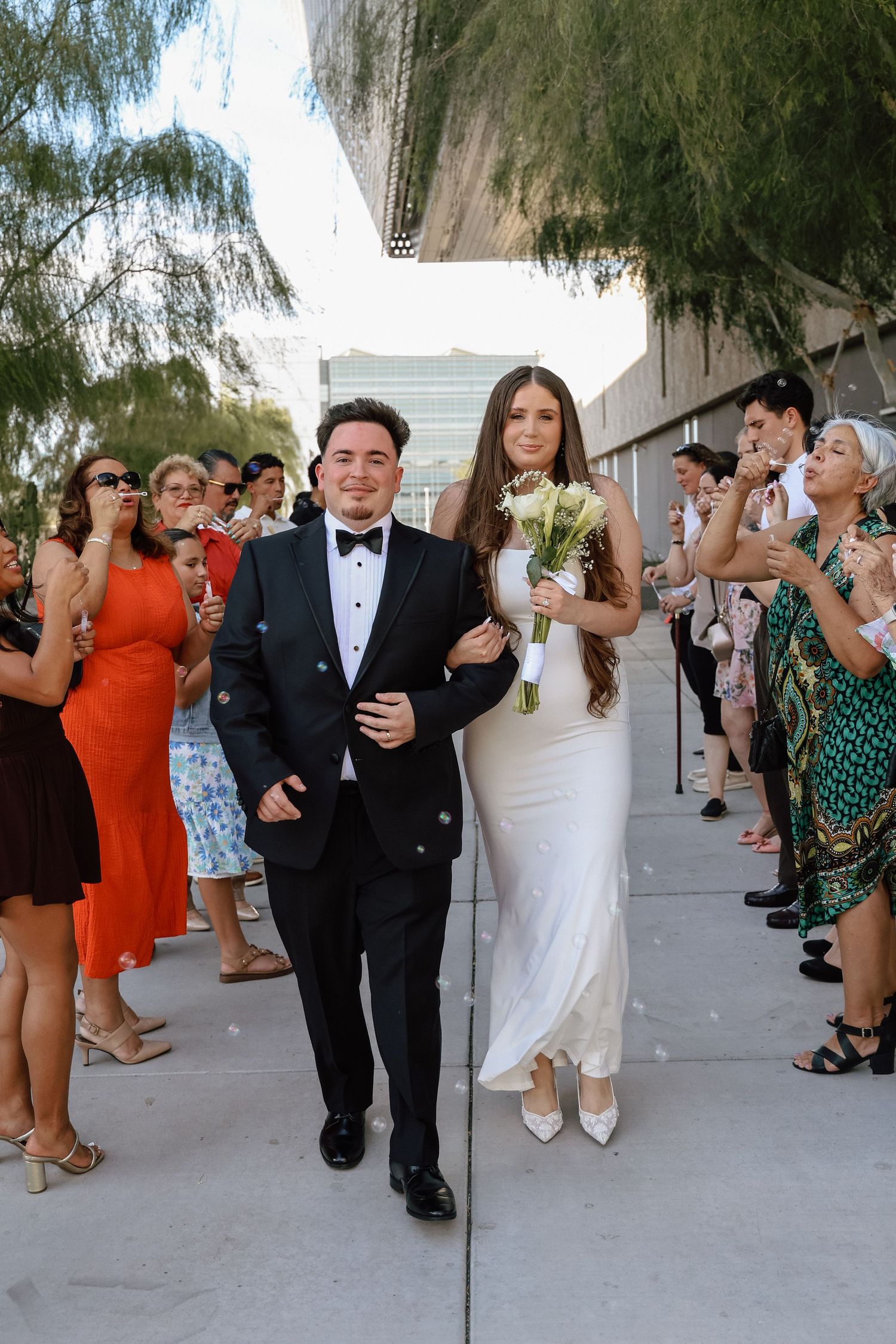 Bride in white gown holding bouquet walks with groom in tuxedo past cheering guests at outdoor wedding ceremony.