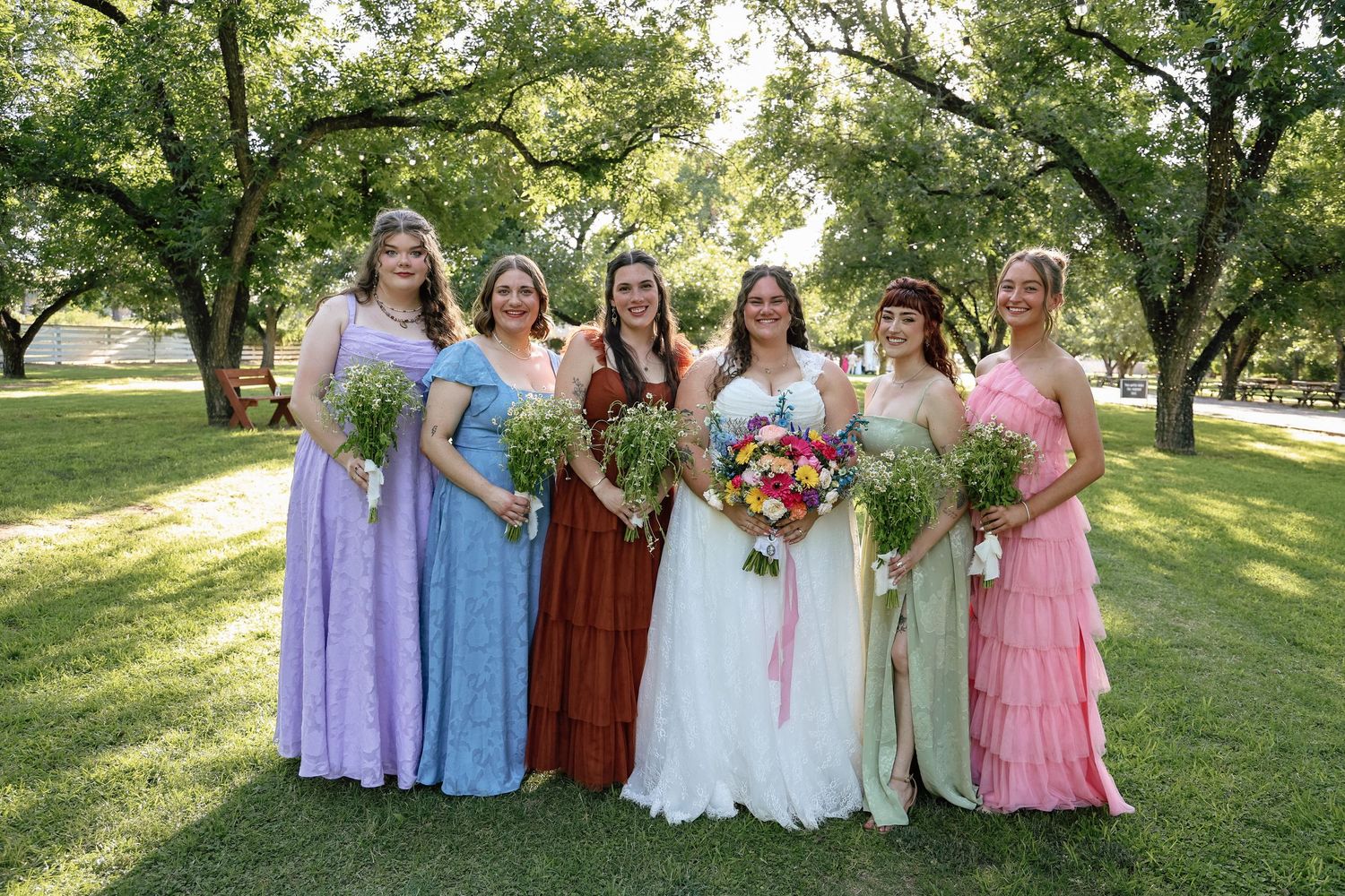 Bride in white gown with five bridesmaids in colorful mismatched dresses holding bouquets at outdoor wedding.