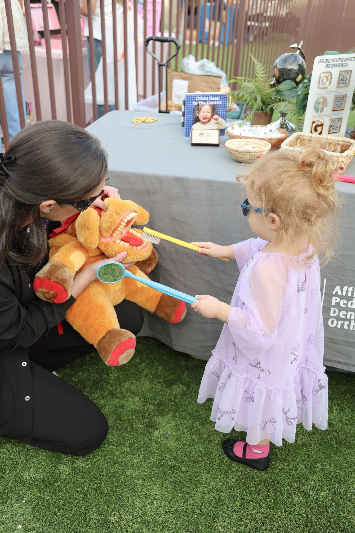A small child in a purple dress plays with a large orange teddy bear on artificial grass.