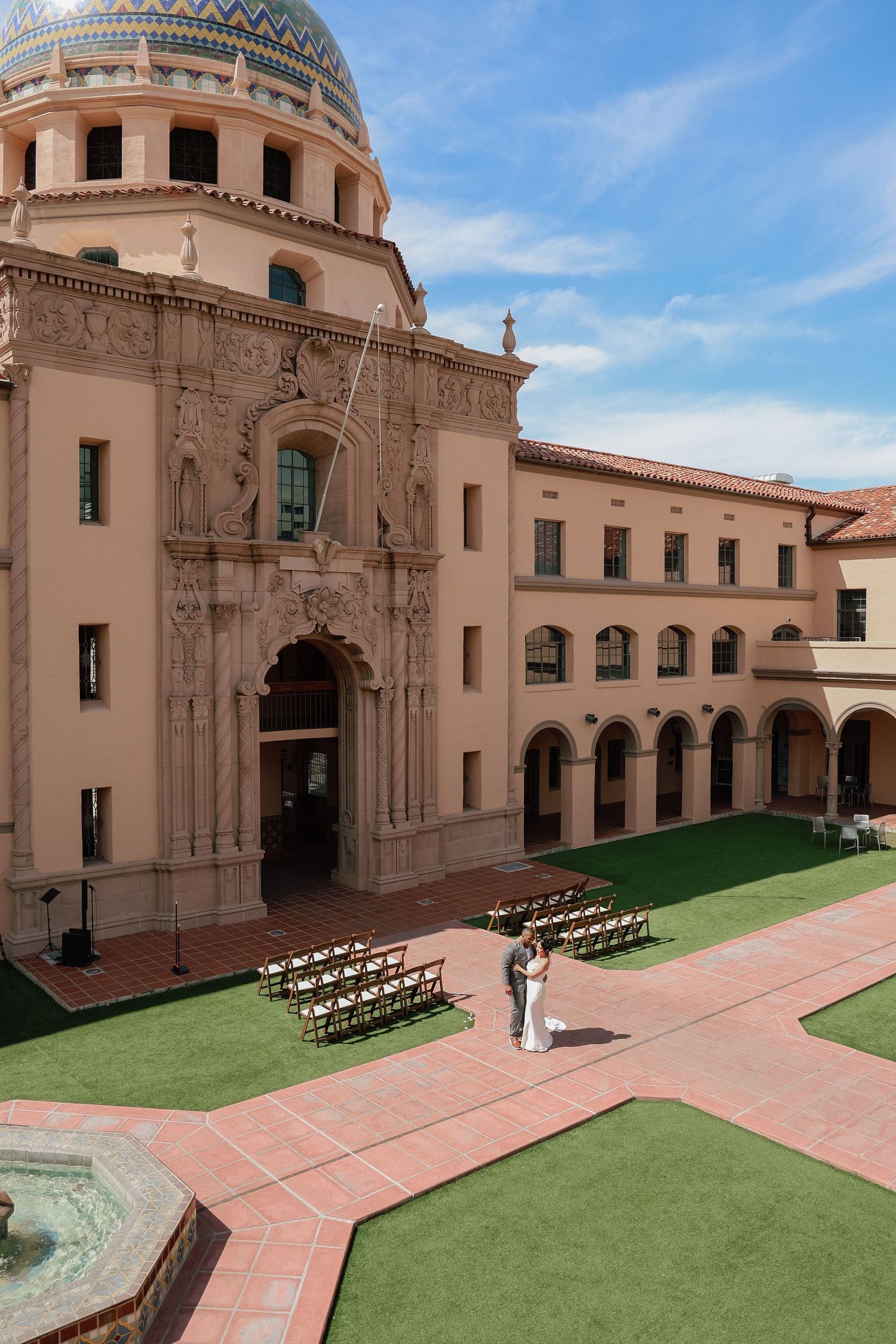 Spanish colonial architecture with domed towers and arched entryways set against blue sky with chairs on lawn.