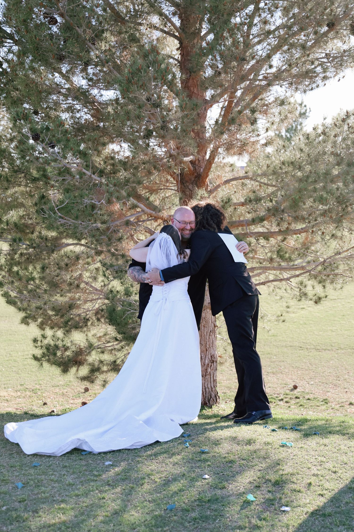 Bride and groom sharing an emotional hug with a guest near a pine tree during an outdoor wedding ceremony.