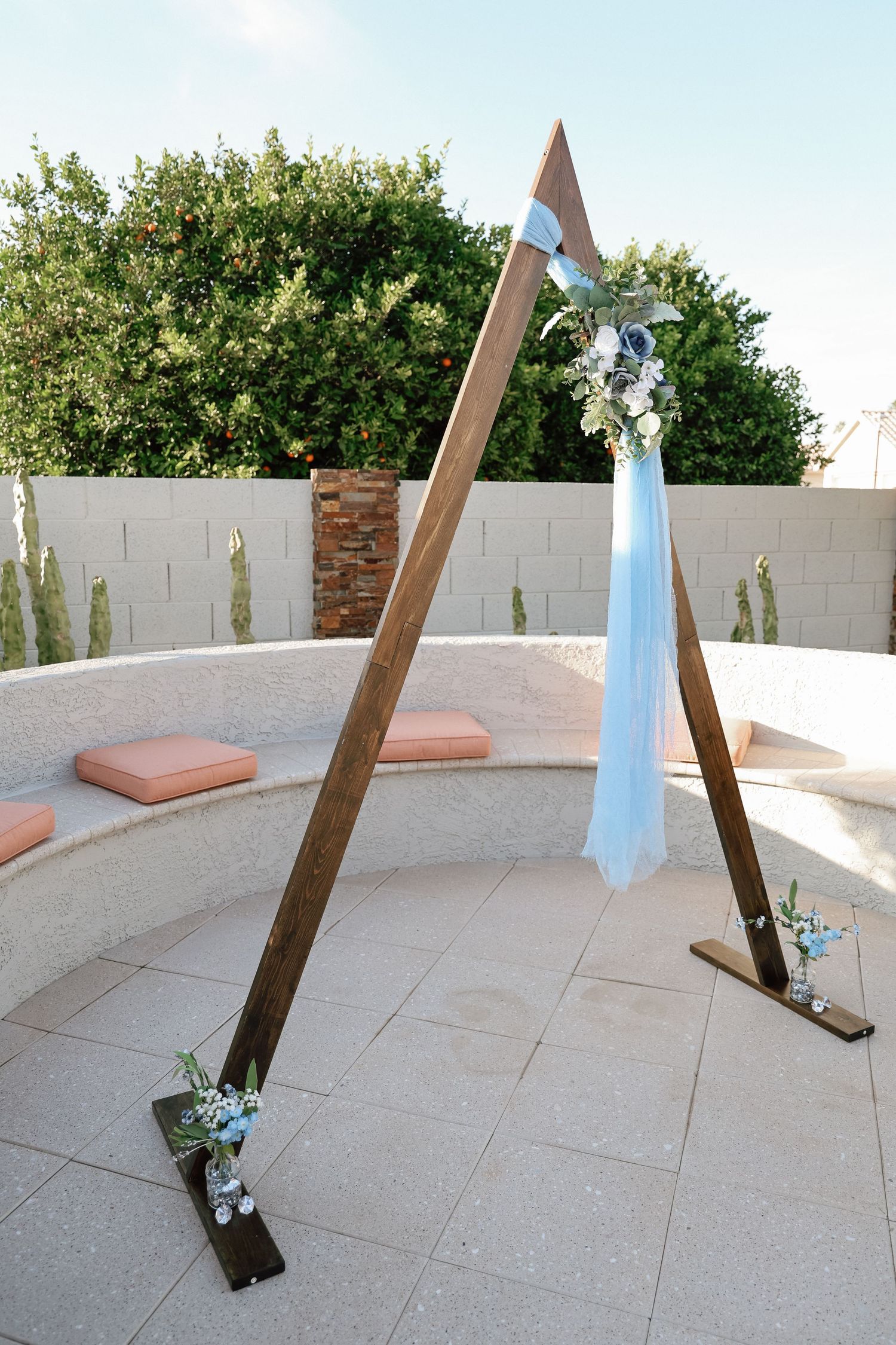 Rustic wooden triangular wedding arch decorated with blue fabric and white flowers in an outdoor patio setting.