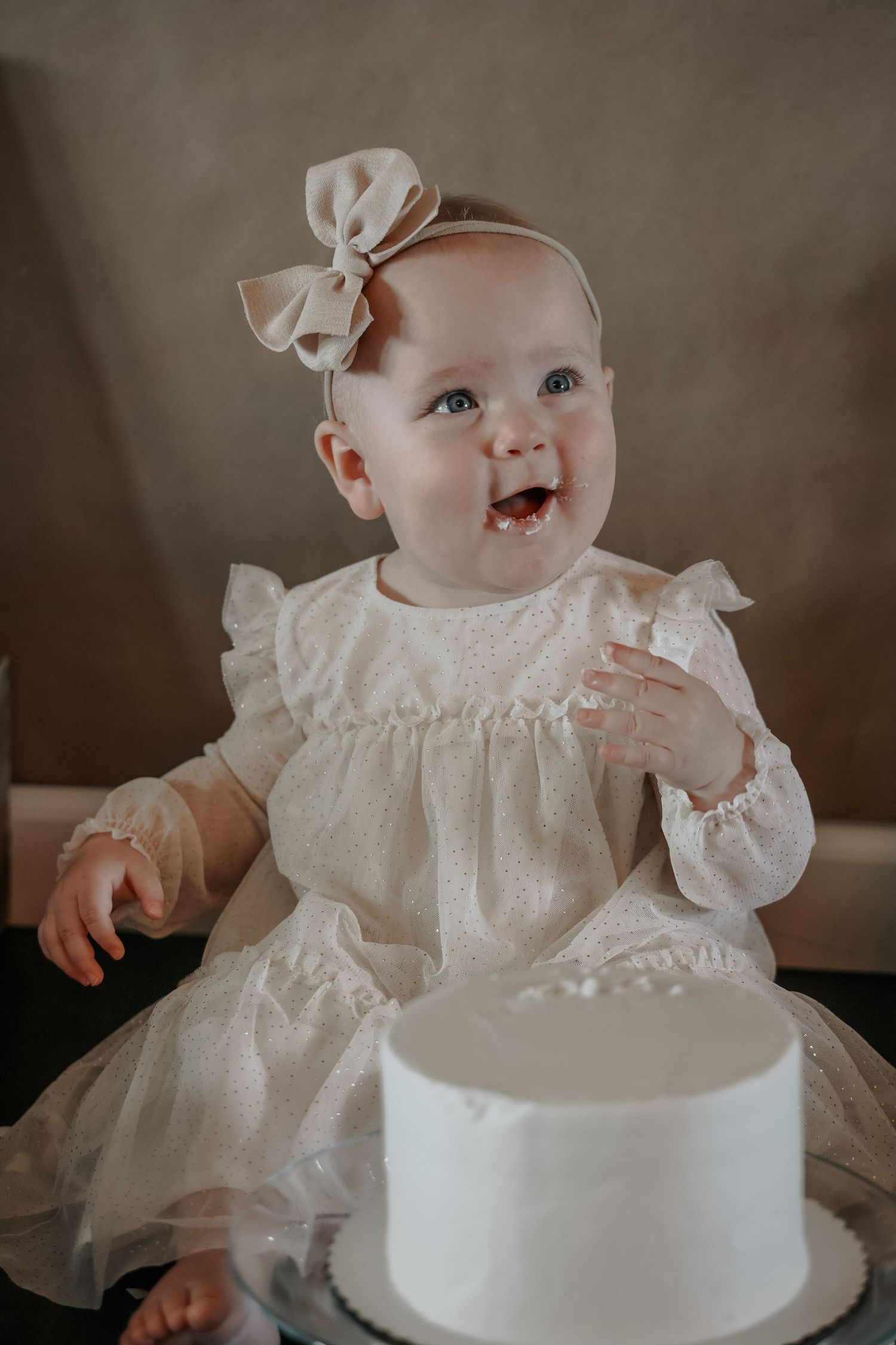 Baby in white lace dress celebrates first birthday with smash cake.
