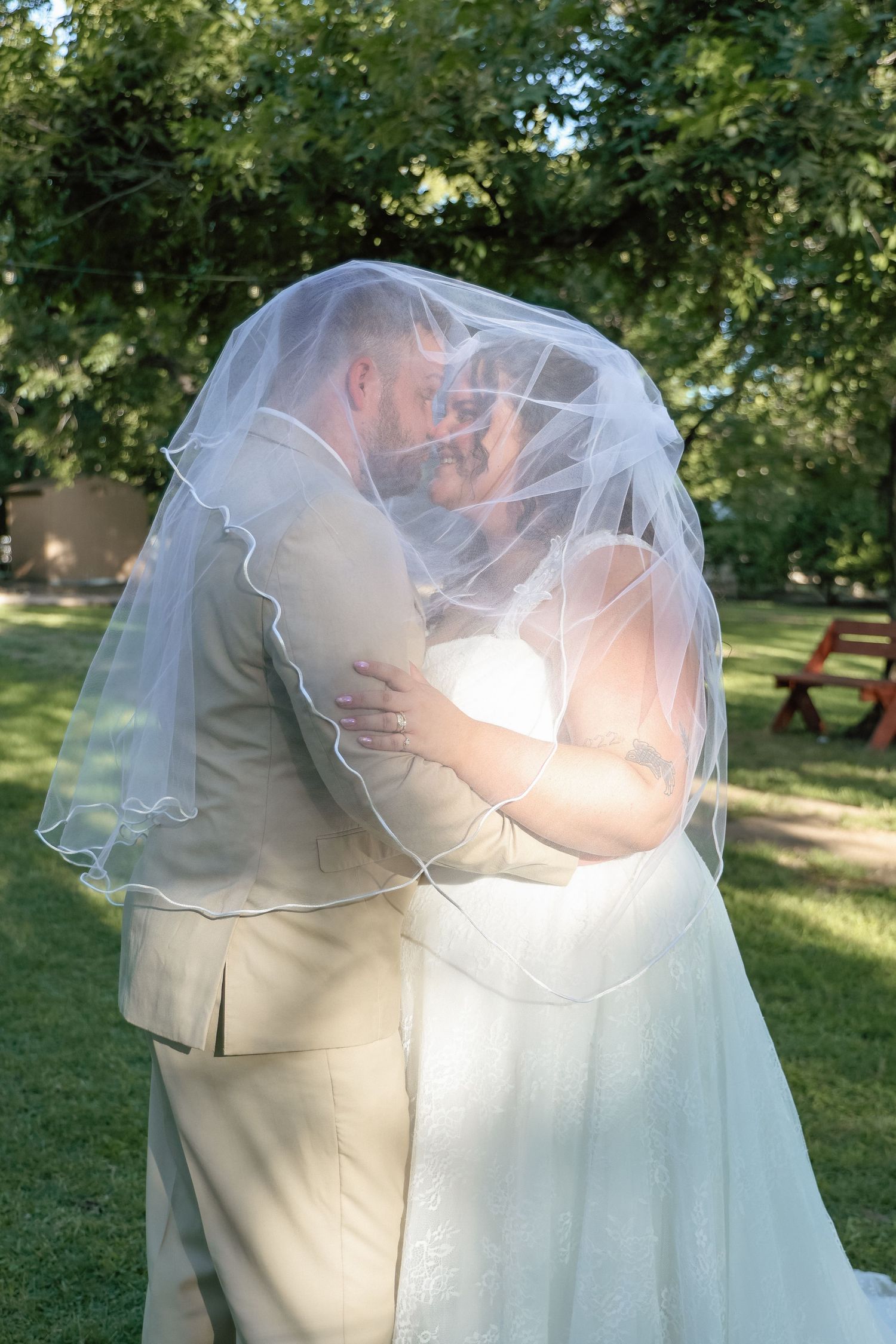Bride and groom share a romantic kiss under a flowing white wedding veil outdoors in a lush green park.