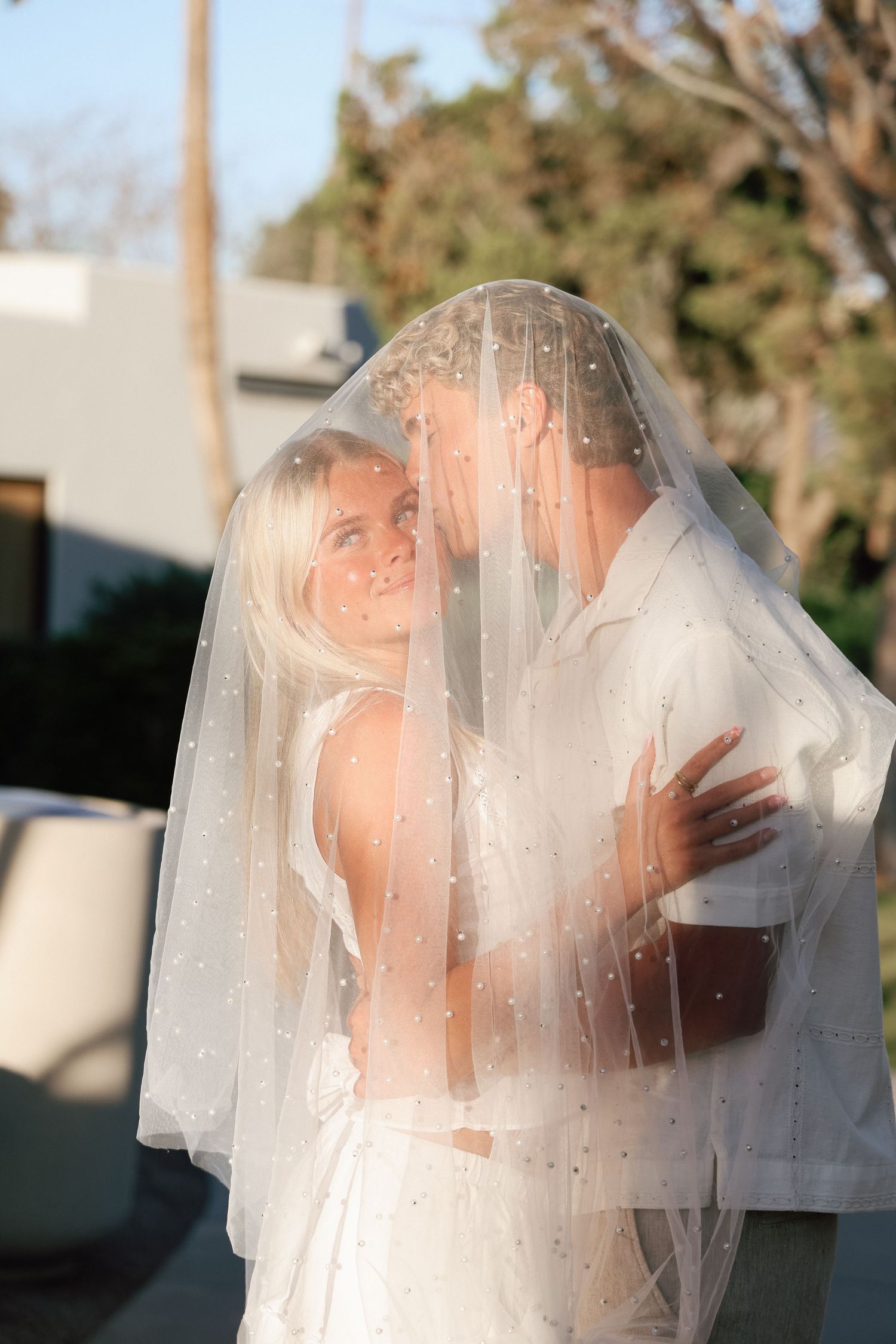 A romantic wedding moment captured through a sheer white veil during sunset with warm golden lighting.
