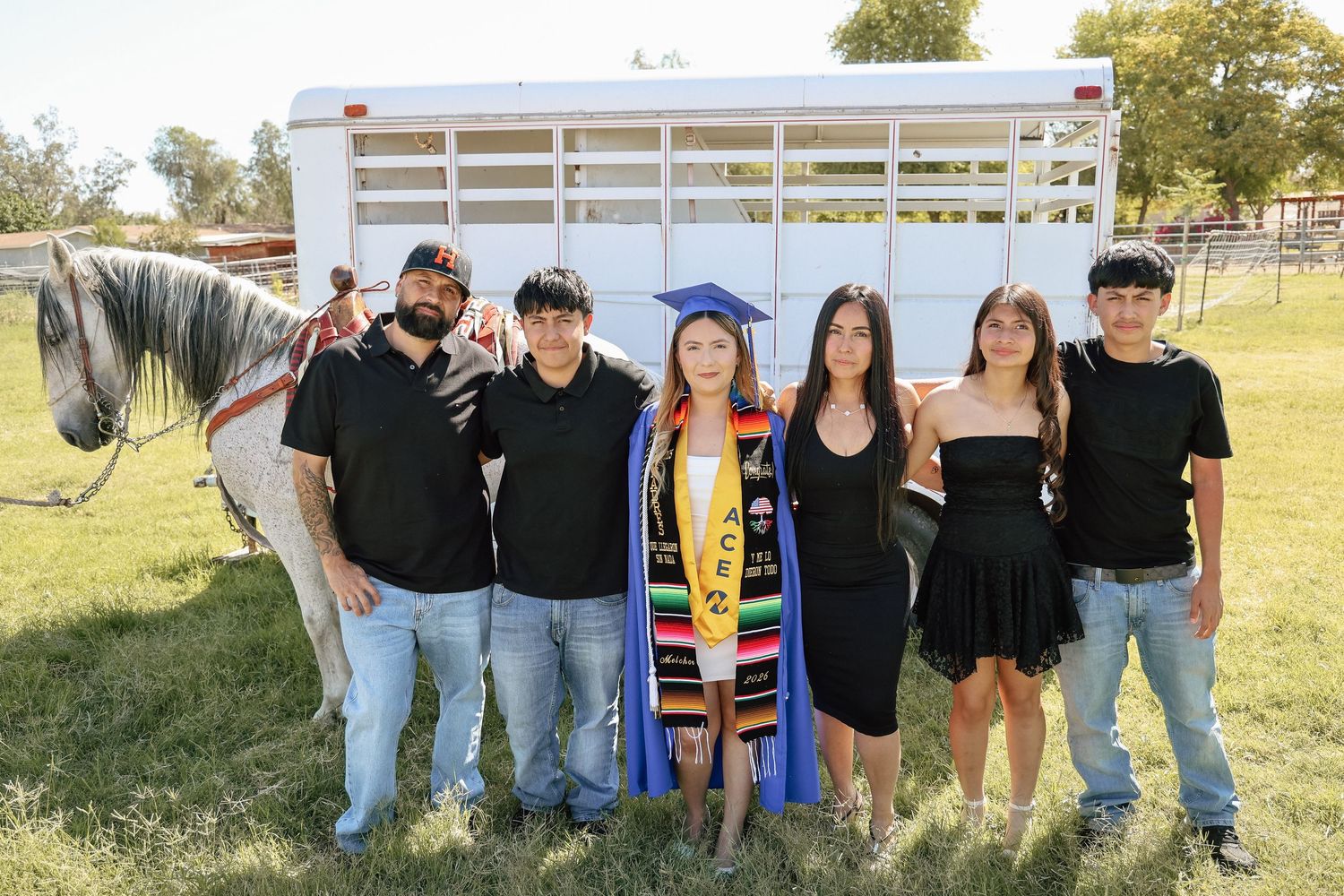 Group of six people posing with a horse at an outdoor graduation celebration, graduate wearing cap and colorful sash.