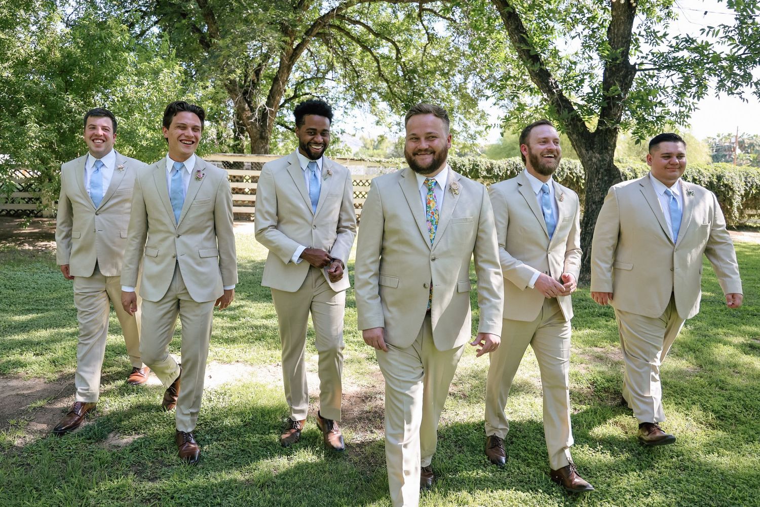 Six groomsmen in matching tan suits with light blue ties walk together outdoors on green grass beneath shady trees.