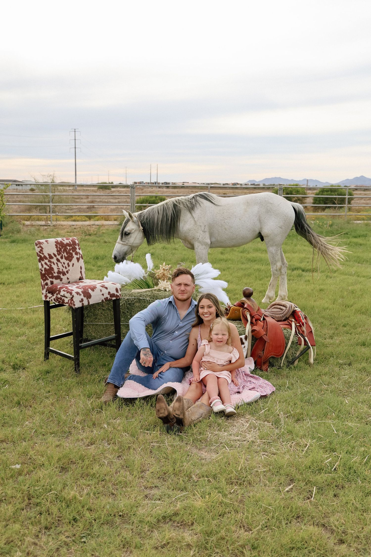 A man and two young girls sit on grass near a gray horse, cow-print chair, and saddle on a farm at dusk.