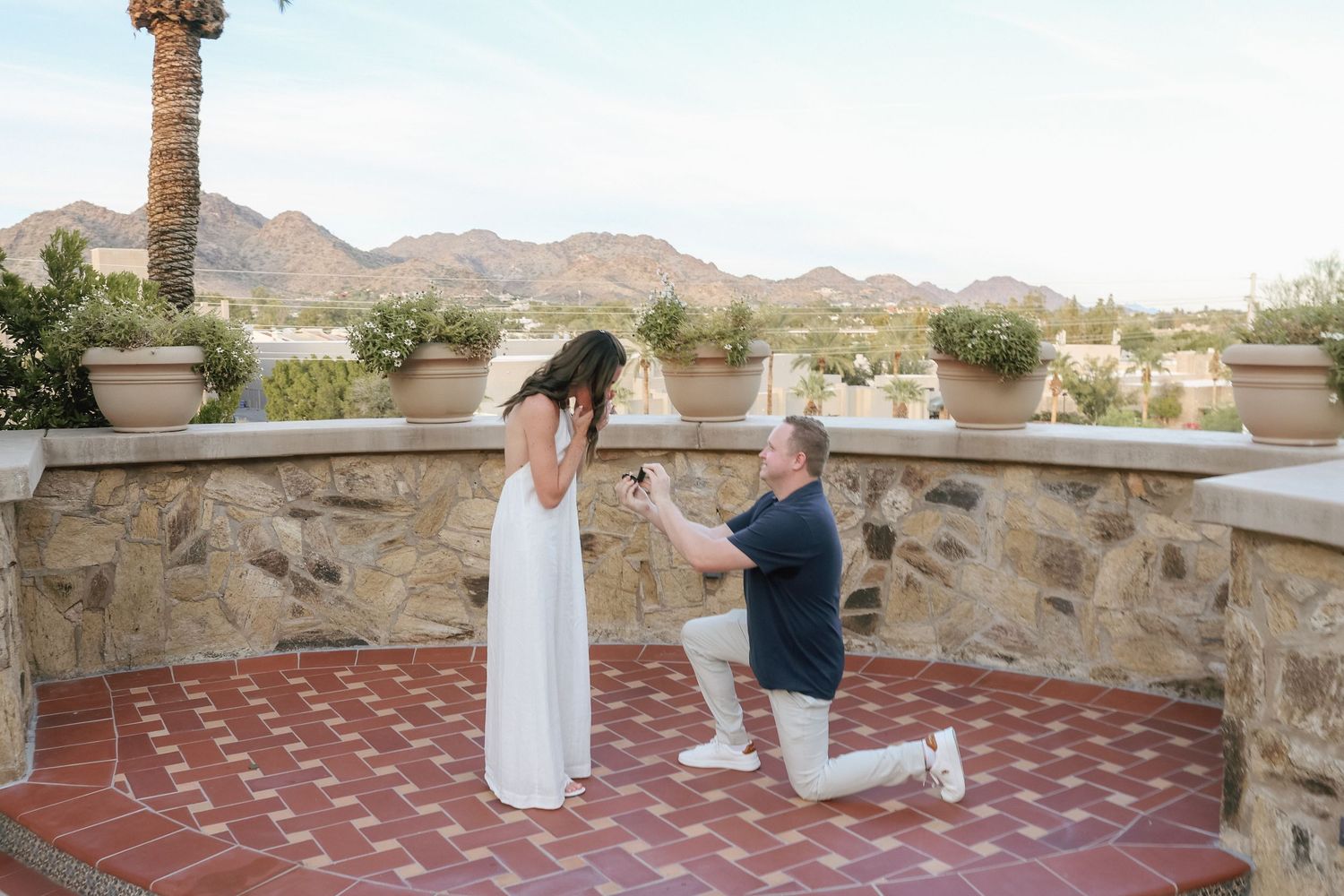 Man proposes to woman on one knee on a scenic terrace with mountain views and lush potted plants in the background.