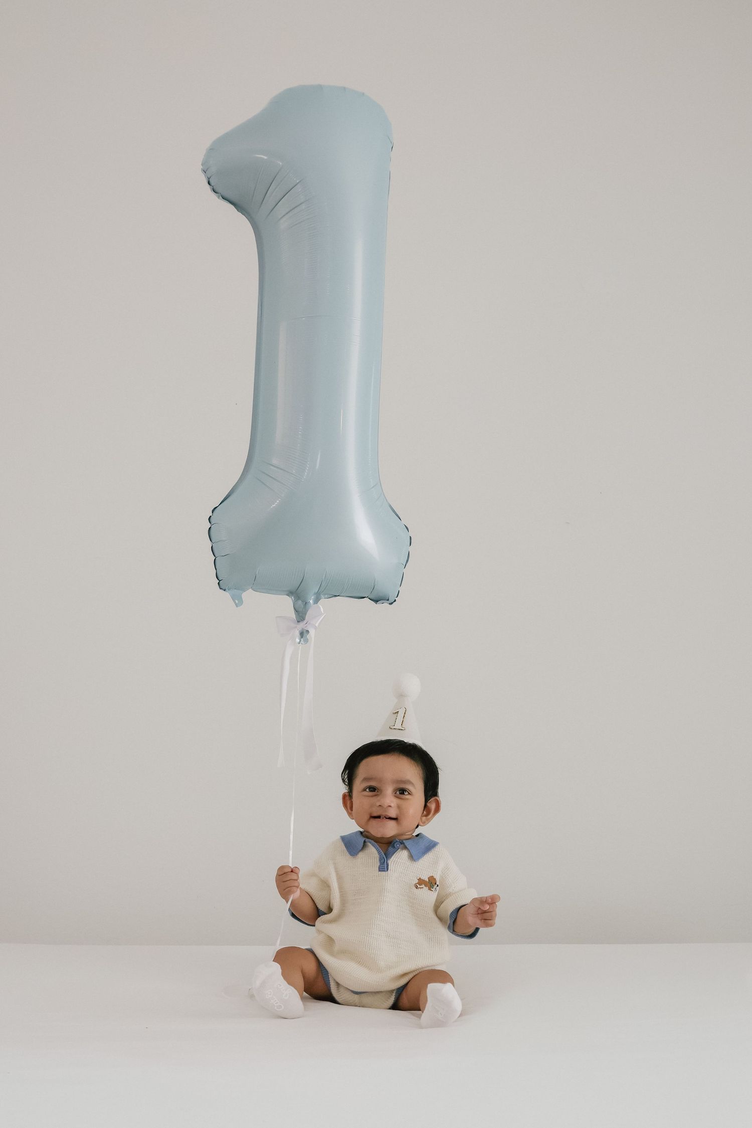 A smiling baby in a cream outfit sits on a white floor holding a light blue number 1 balloon for their first birthday.