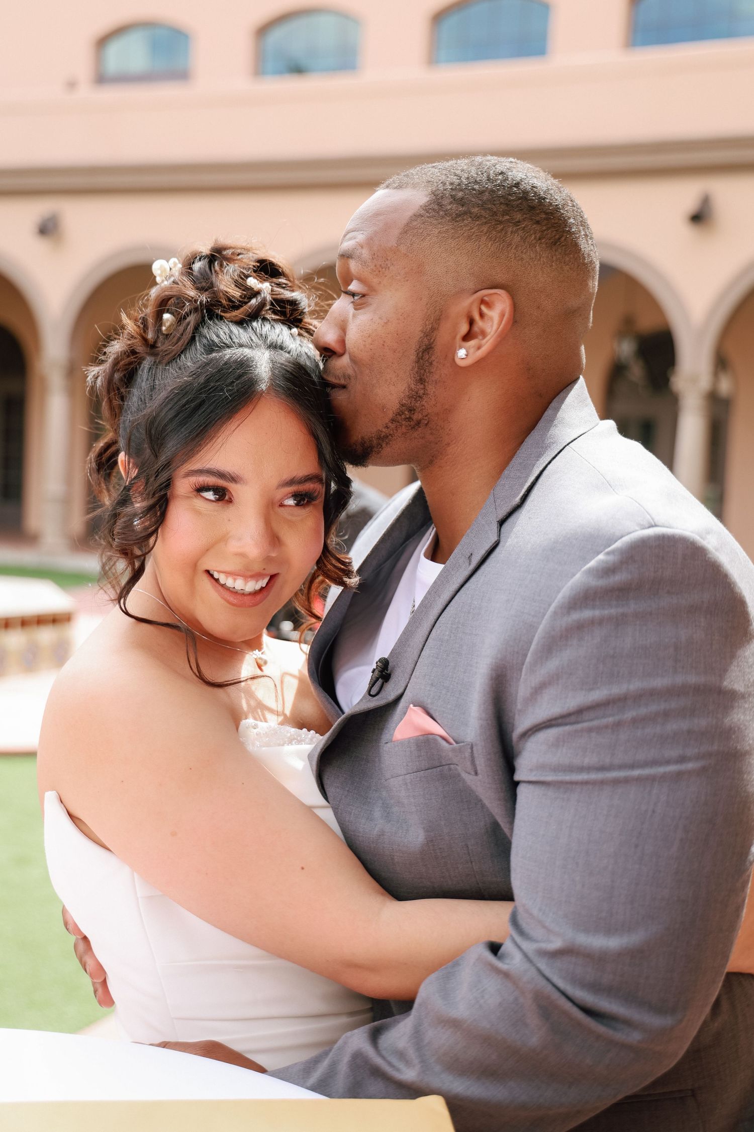 A groom in a gray suit kisses his smiling bride in a white dress during their wedding celebration outdoors.