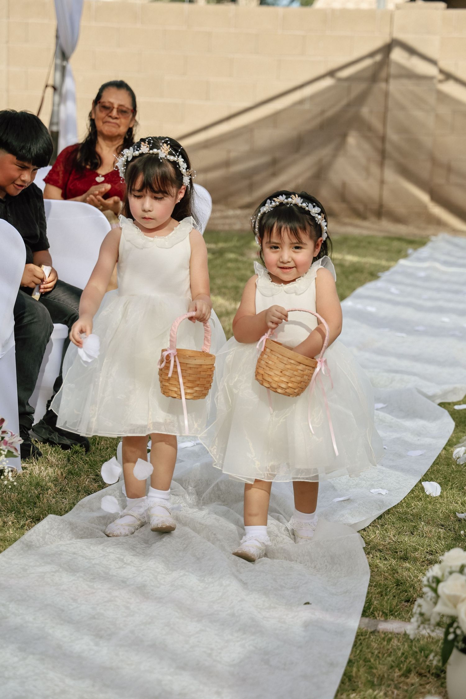 Two adorable flower girls in white dresses and floral headbands walk down the aisle carrying baskets at an outdoor wedding.