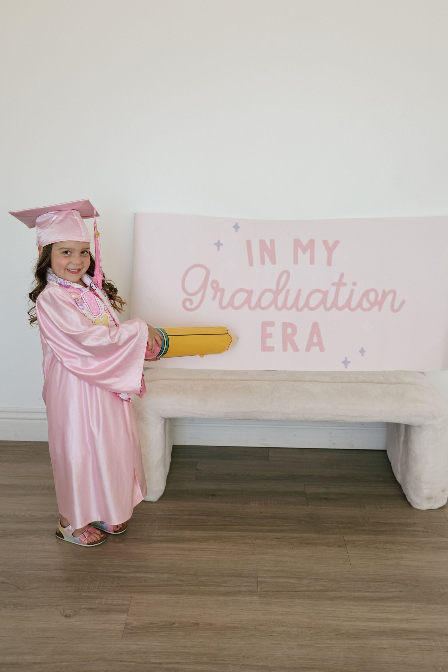 Young girl in pink graduation cap and gown posing beside a bench with an In My Graduation Era sign.