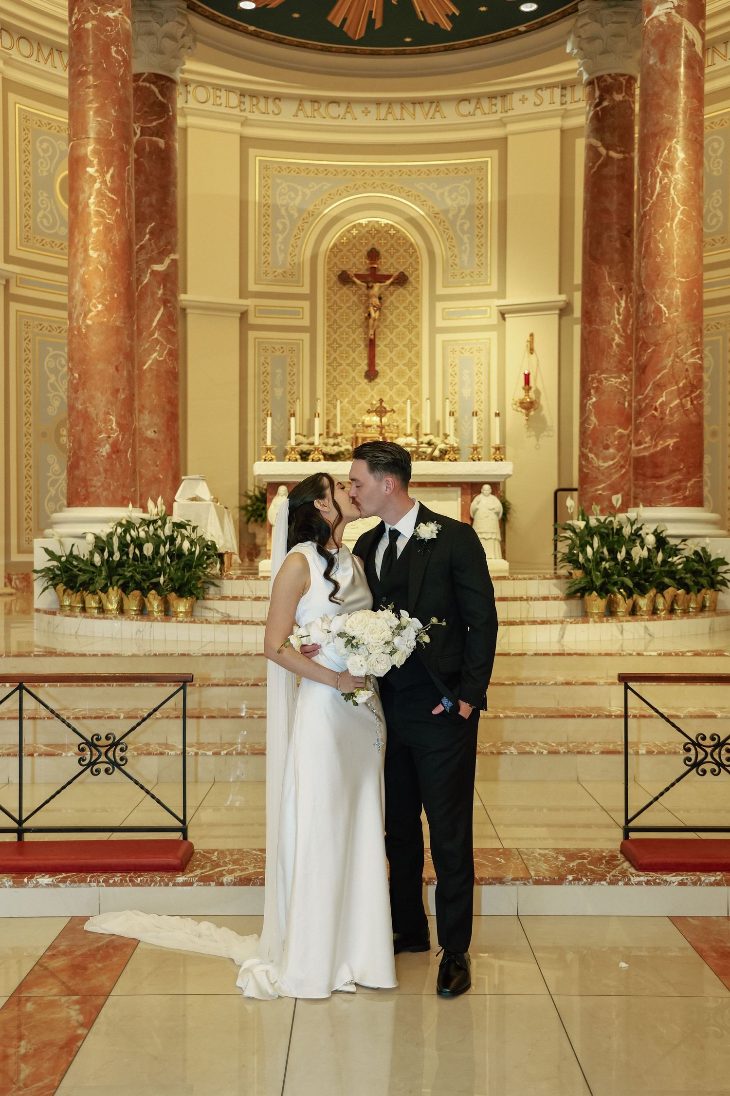 Bride and groom share a romantic moment at ornate church altar, bride holding white bouquet in elegant wedding gown.