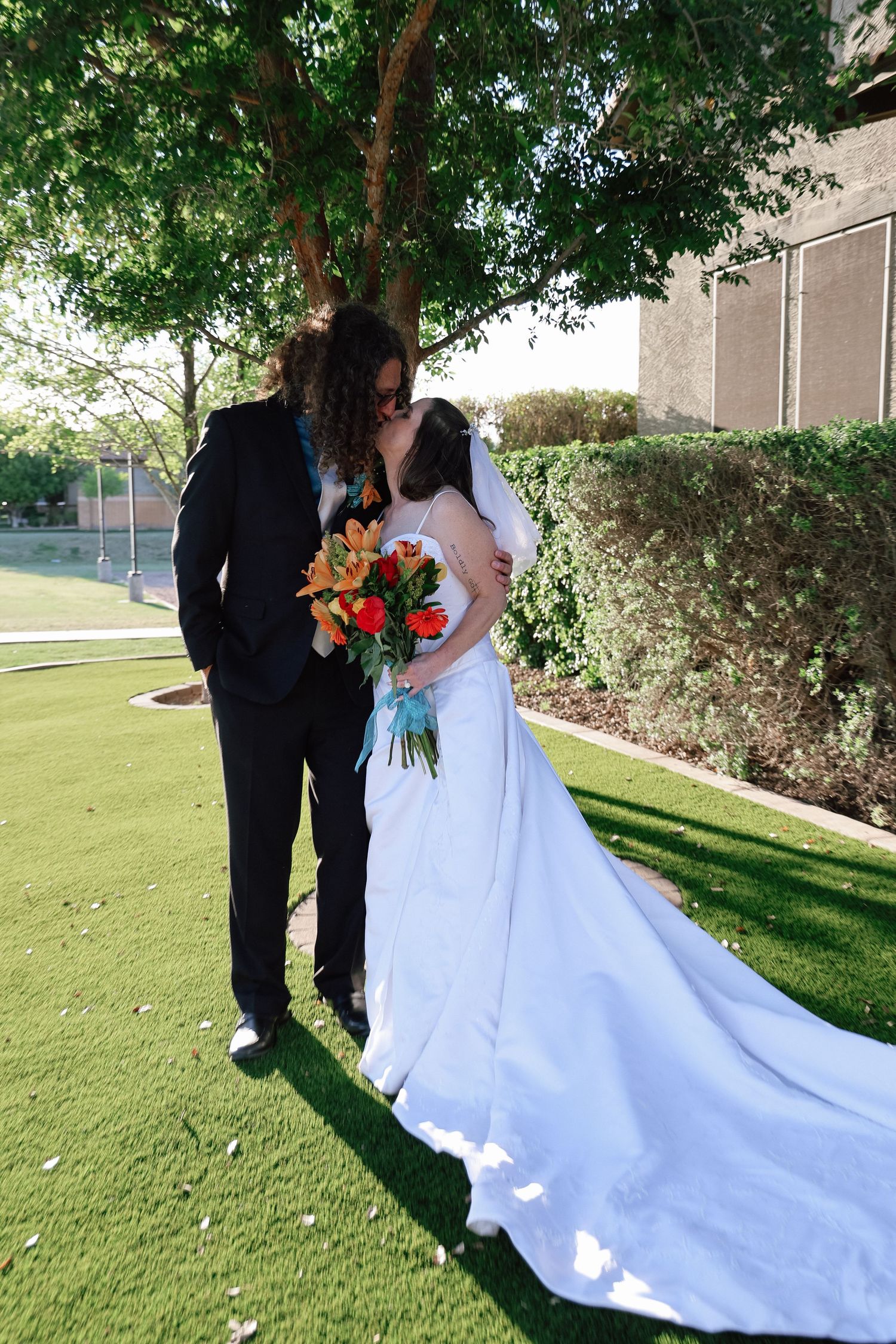 Newlywed couple sharing a kiss outdoors, bride in white gown holding red bouquet, groom in black suit under green trees.