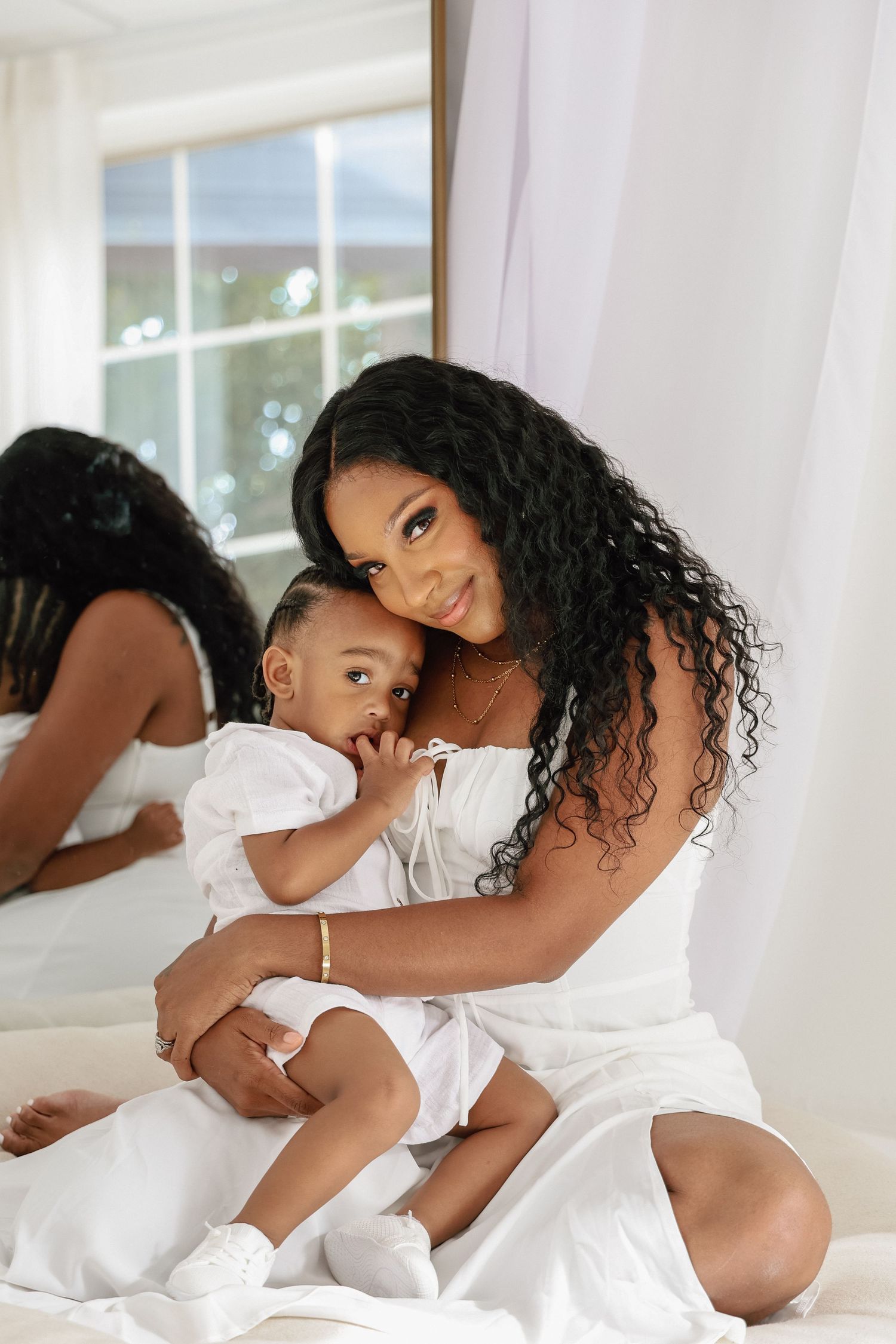 A young Black mother in a white dress holds her baby on a bed, both dressed in white, with a mirror reflection behind them.