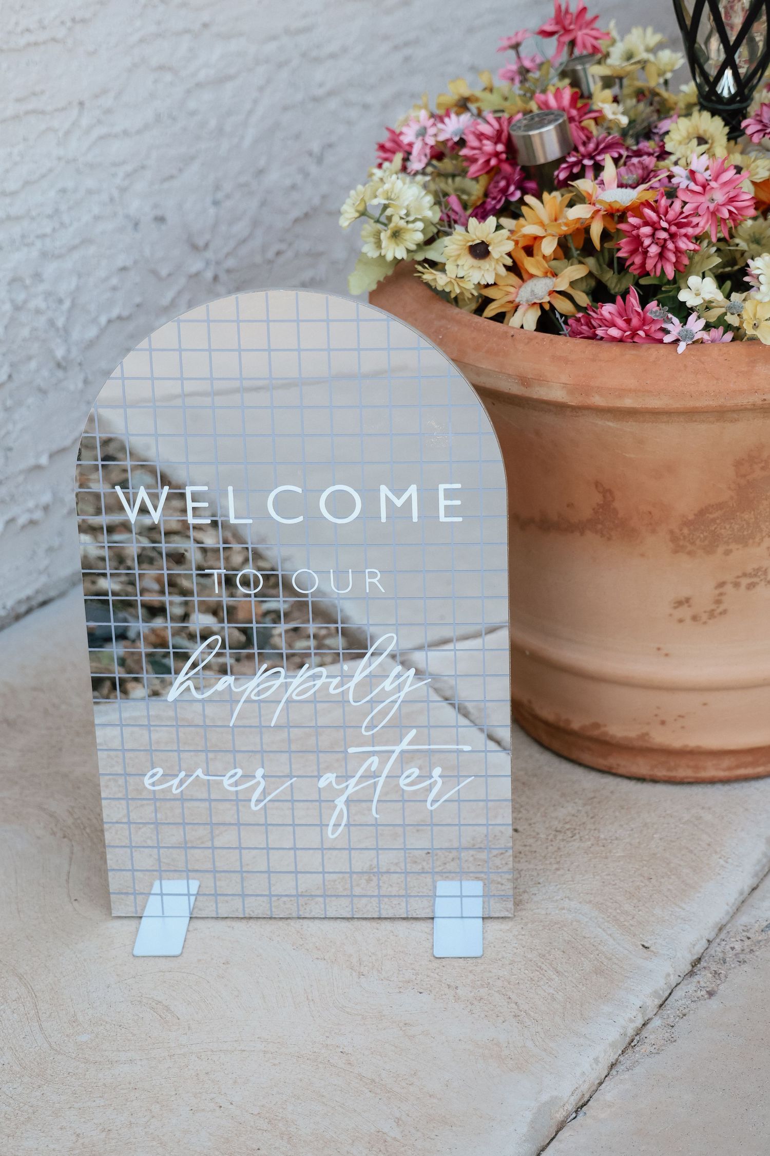 Acrylic wedding welcome sign reading happily ever after, displayed on stone steps beside a terra cotta pot of colorful flowers.