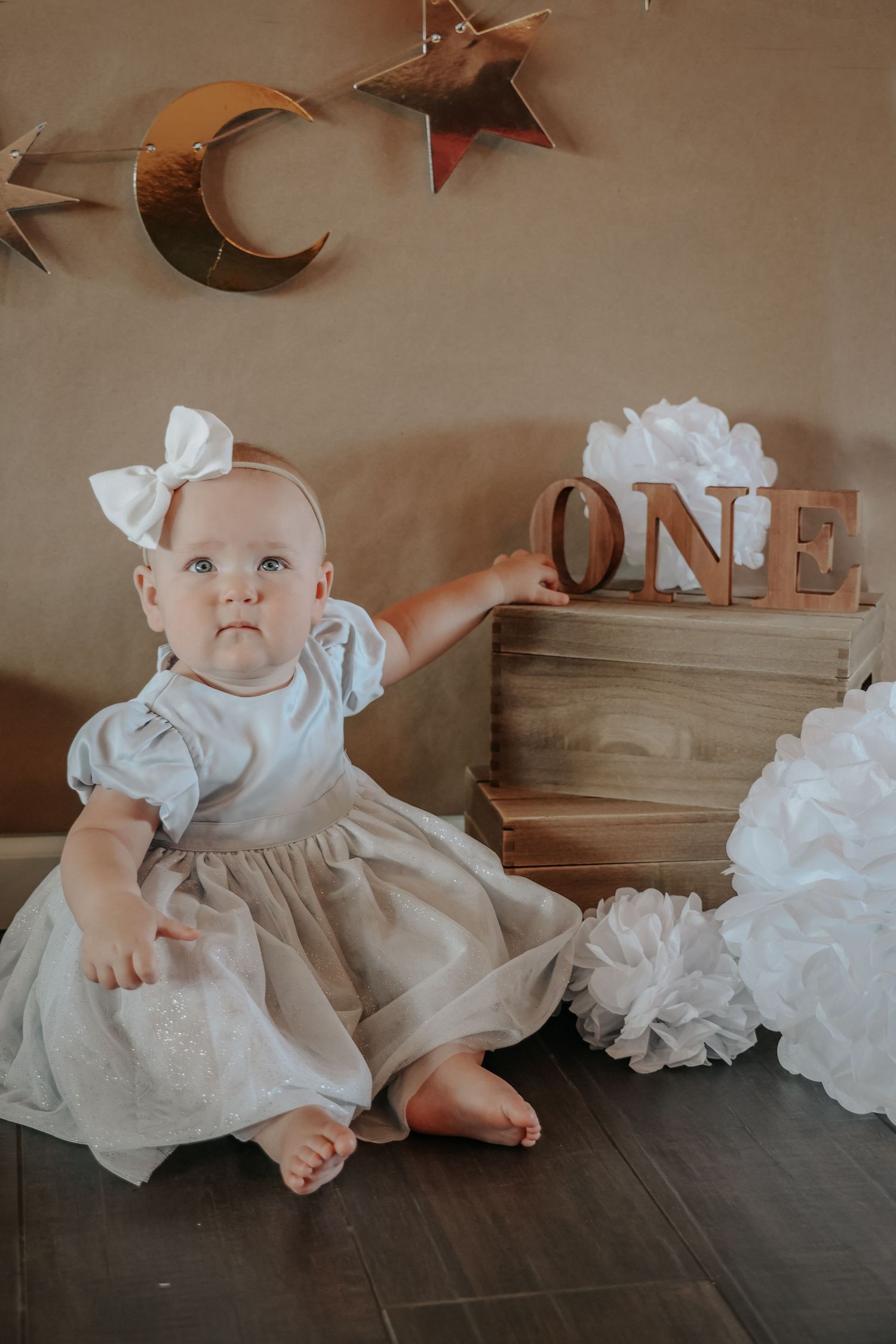 Baby in white dress sitting near wooden ONE sign for first birthday photos.