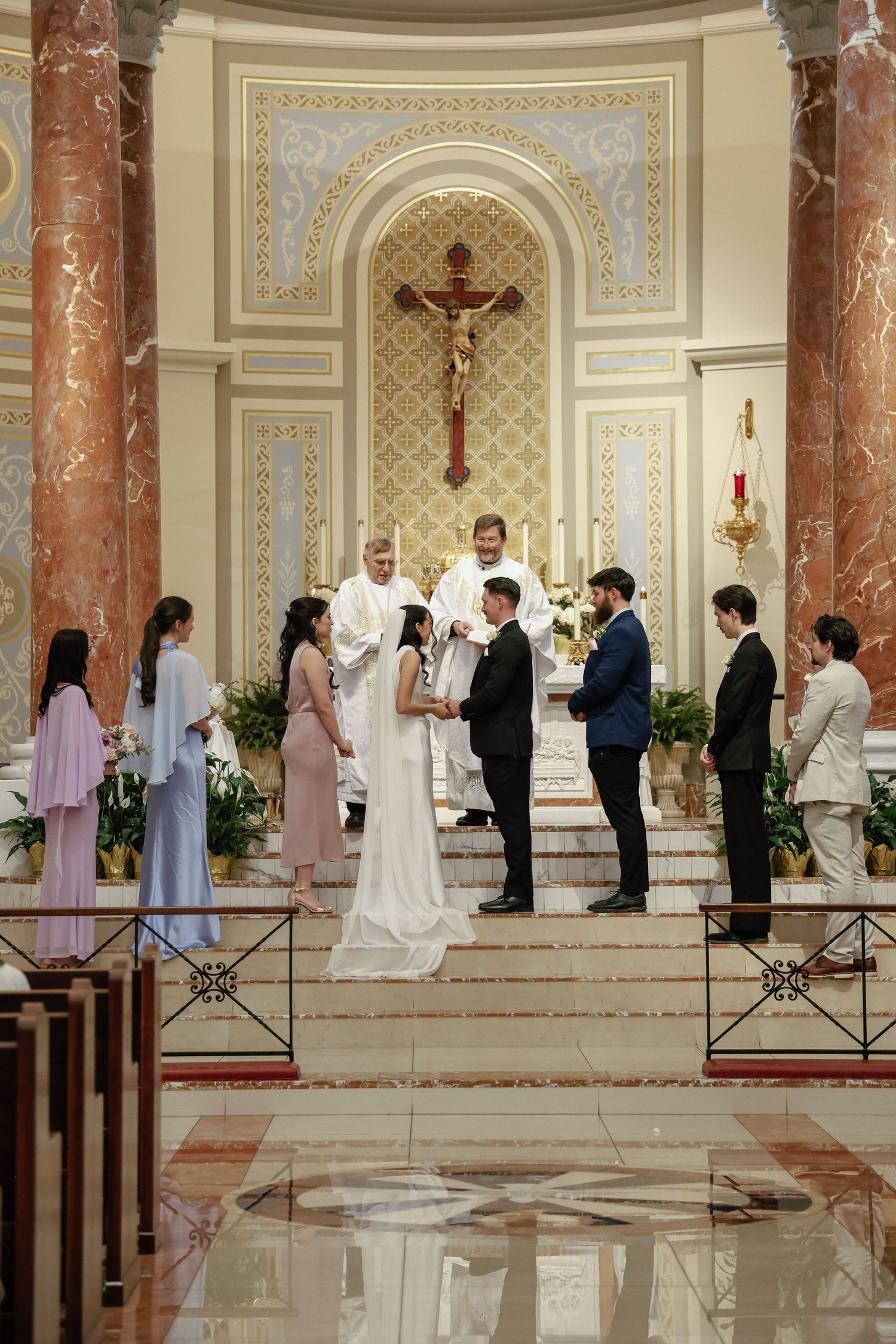 Catholic church wedding ceremony with bride and groom exchanging vows before a priest at an ornate altar with crucifix.