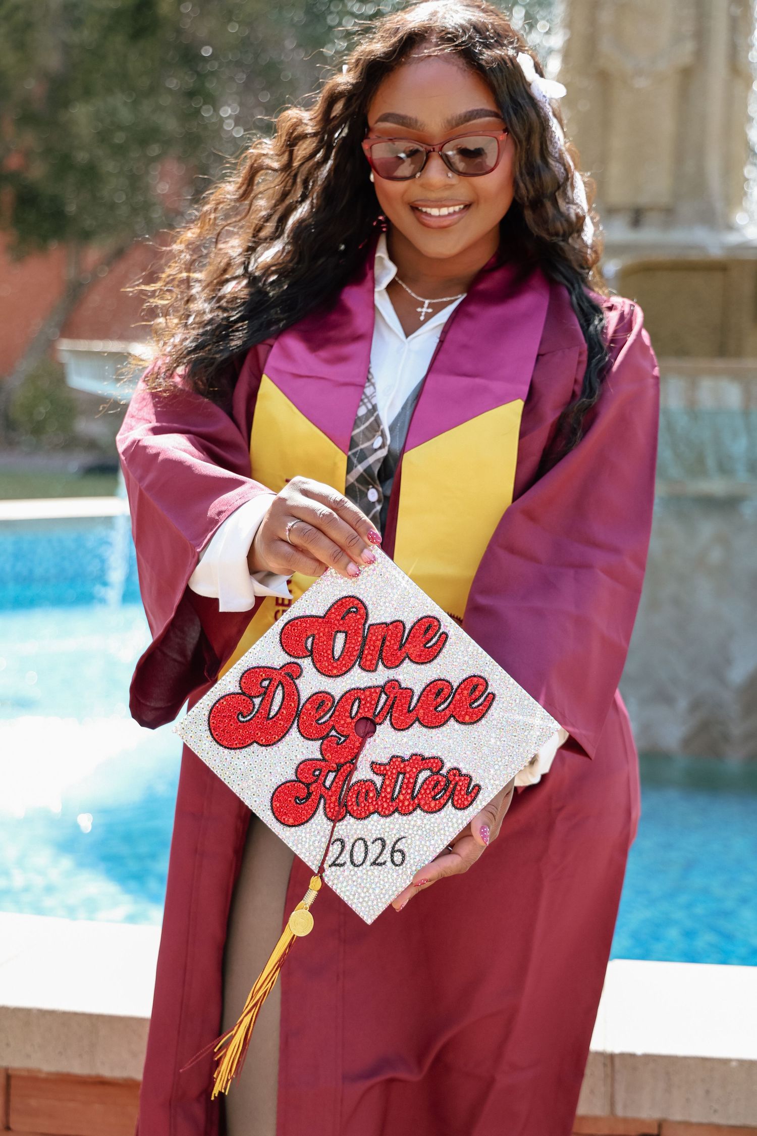 Graduate in maroon and gold cap and gown holds decorated mortarboard reading One Degree Hotter 2026.