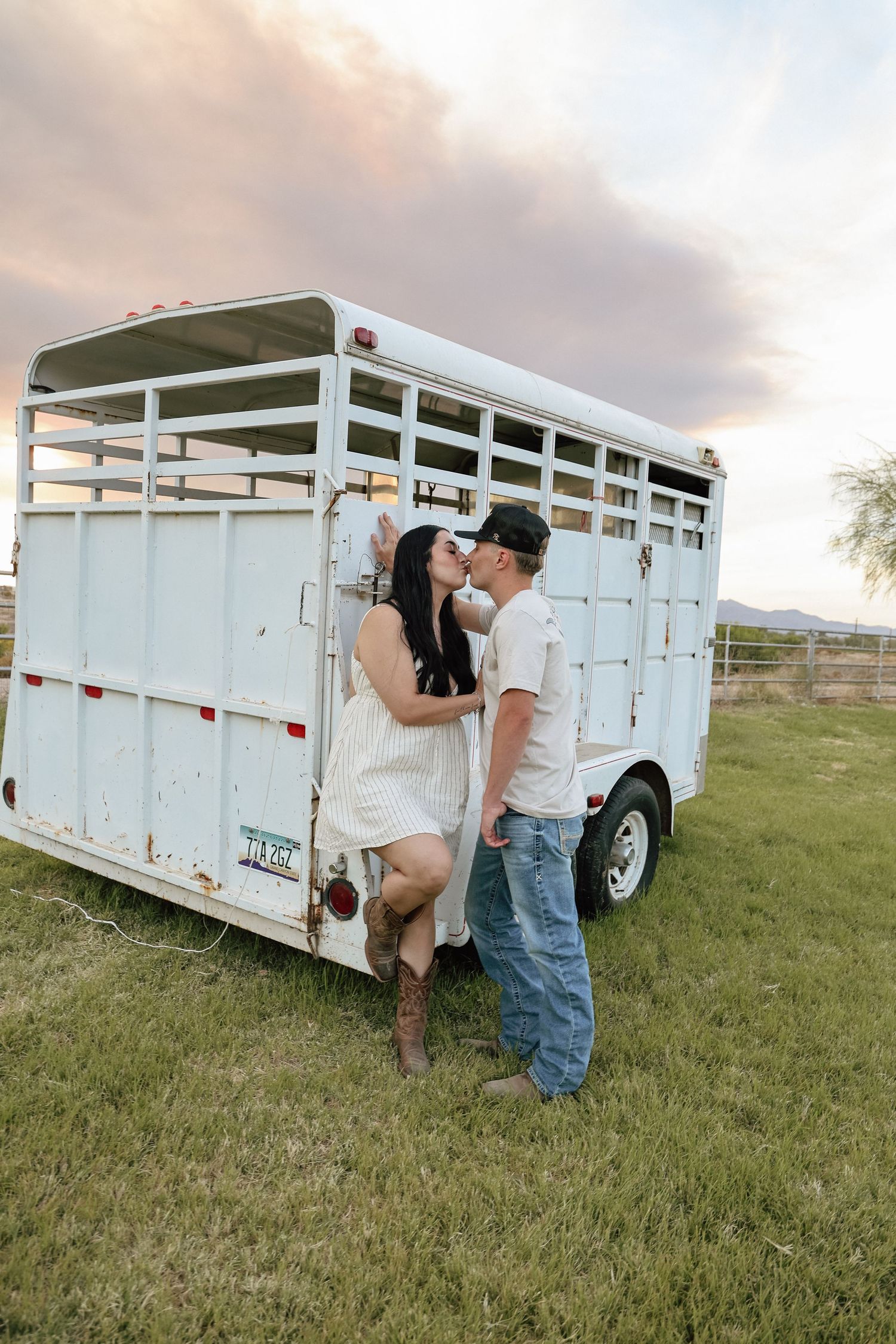 Couple kissing against a white livestock trailer on a green field at sunset, woman in white dress and cowboy boots.