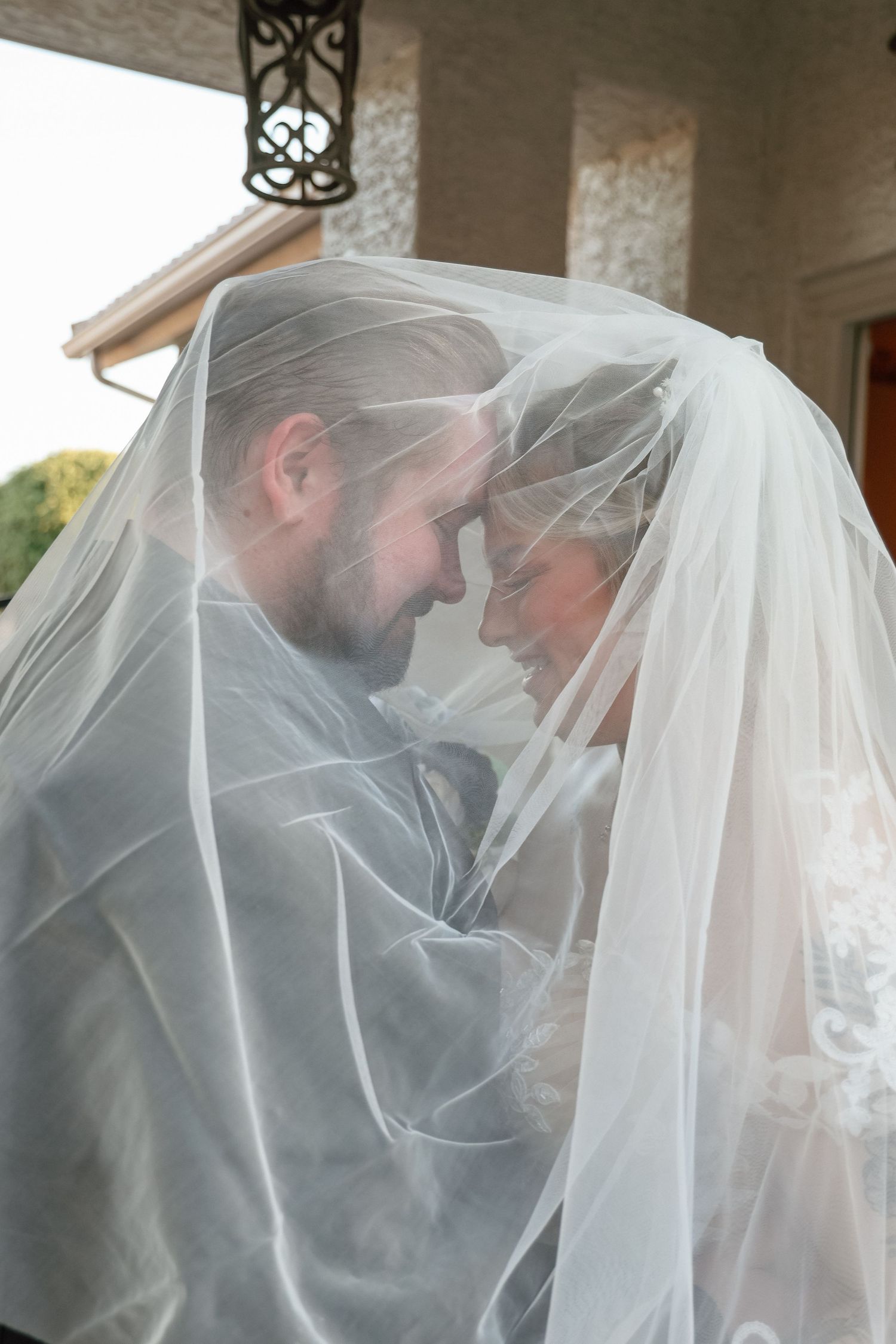 Bride and groom sharing an intimate moment under a flowing white wedding veil, foreheads touching tenderly.