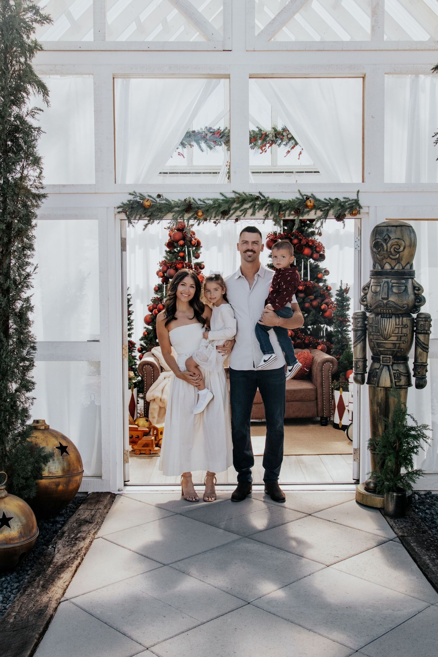 Family holiday photos in white room decorated with Christmas trees and greenery.