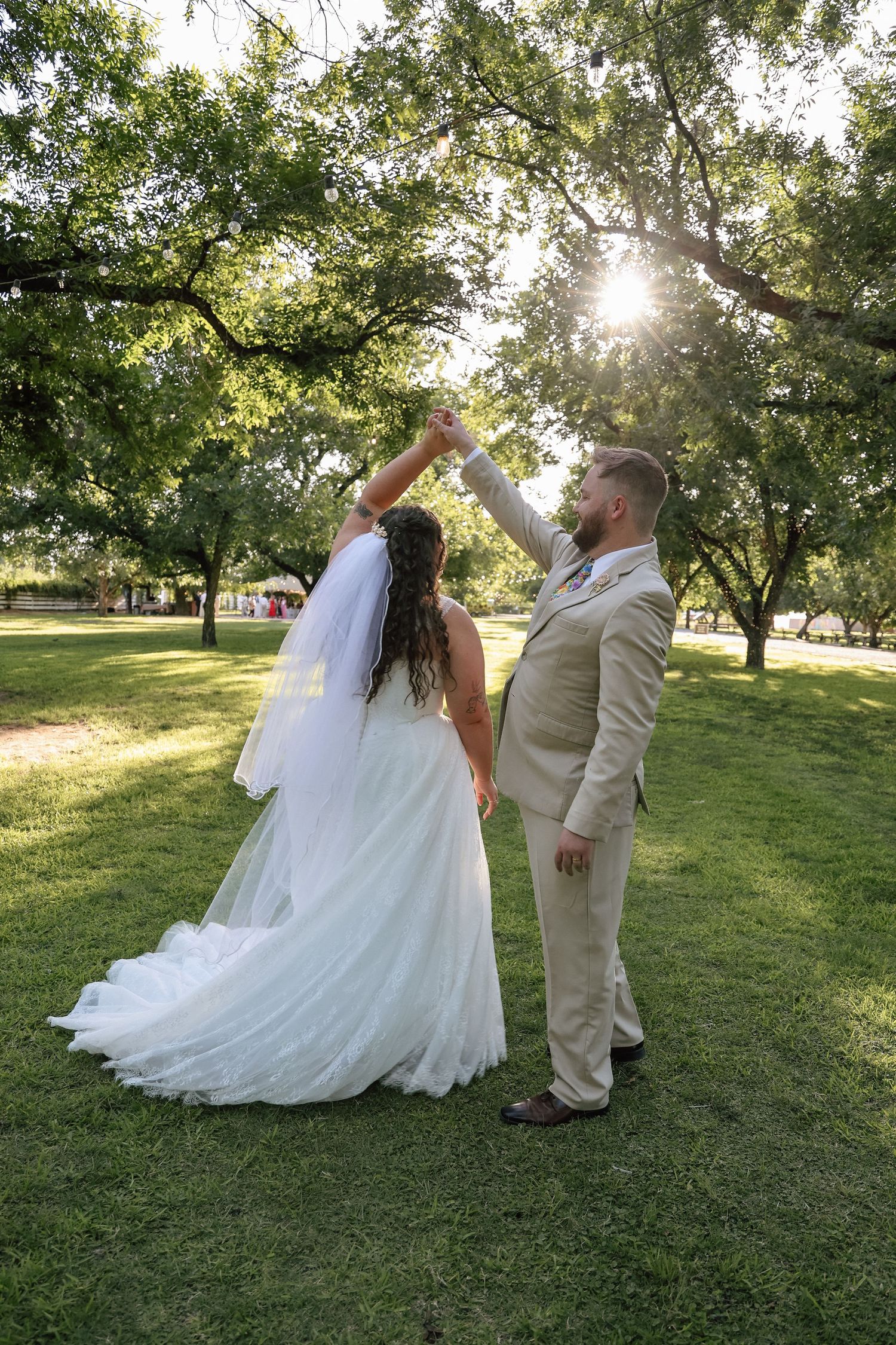 Bride and groom dancing in sunlit park, groom twirling bride in white gown and veil.