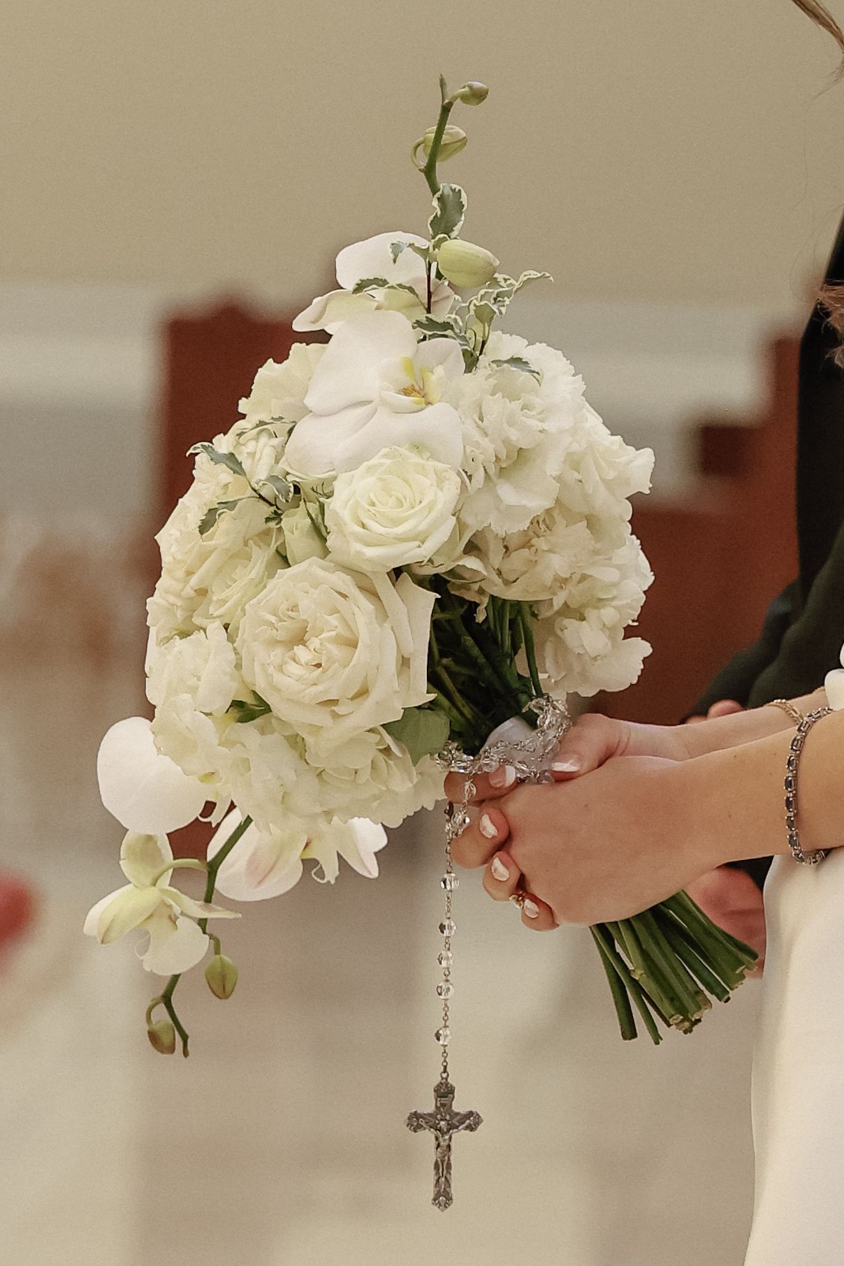 Bride holding white rose and orchid wedding bouquet with silver rosary and crucifix at church ceremony.